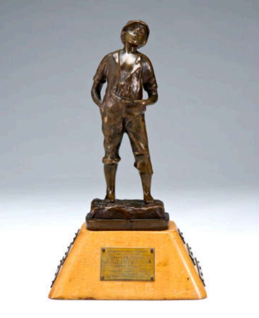 Close-up view of the Whistler Boy Trophy showing the bronze figure of a standing boy in early 20th-century work clothes atop a maple pedestal with engraved brass dedication plaque