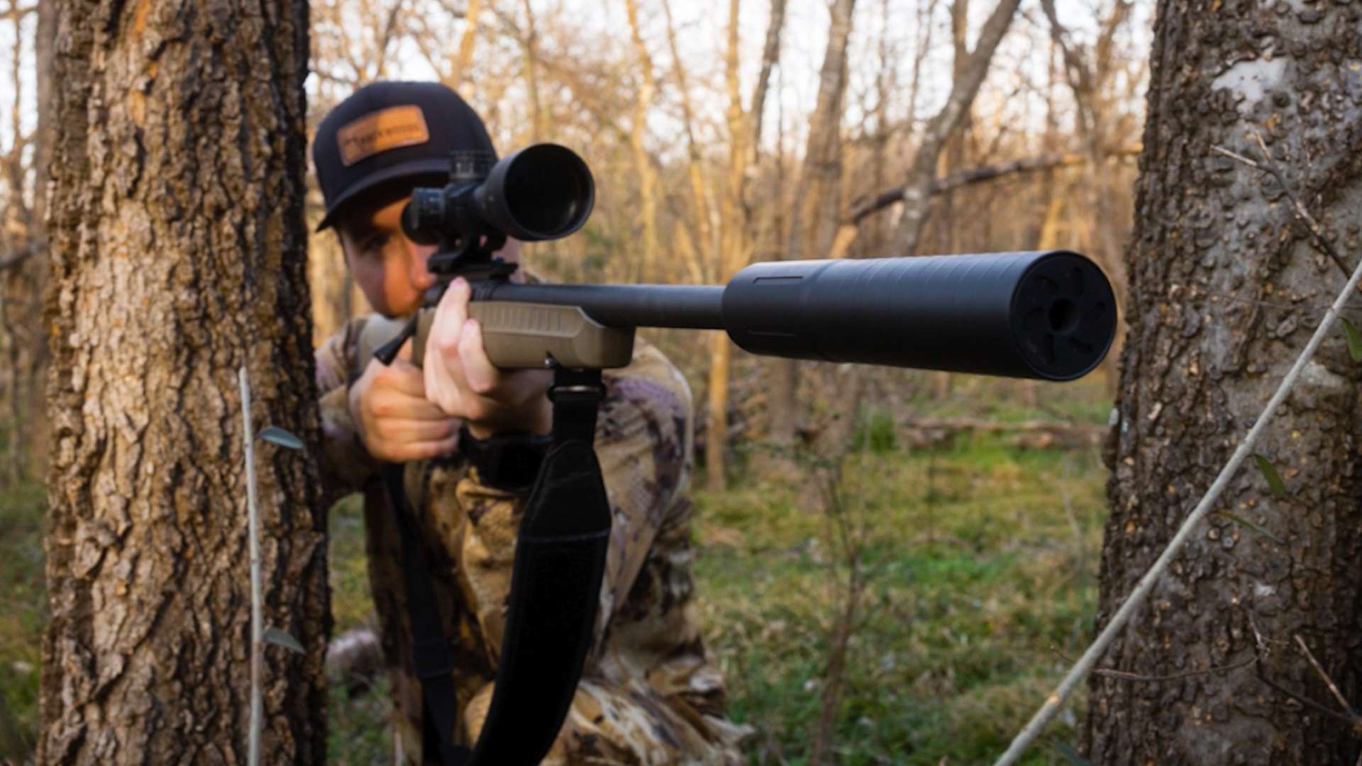 Camouflaged hunter aiming a scoped bolt-action rifle equipped with a Backwoods suppressor between two trees in a wooded area