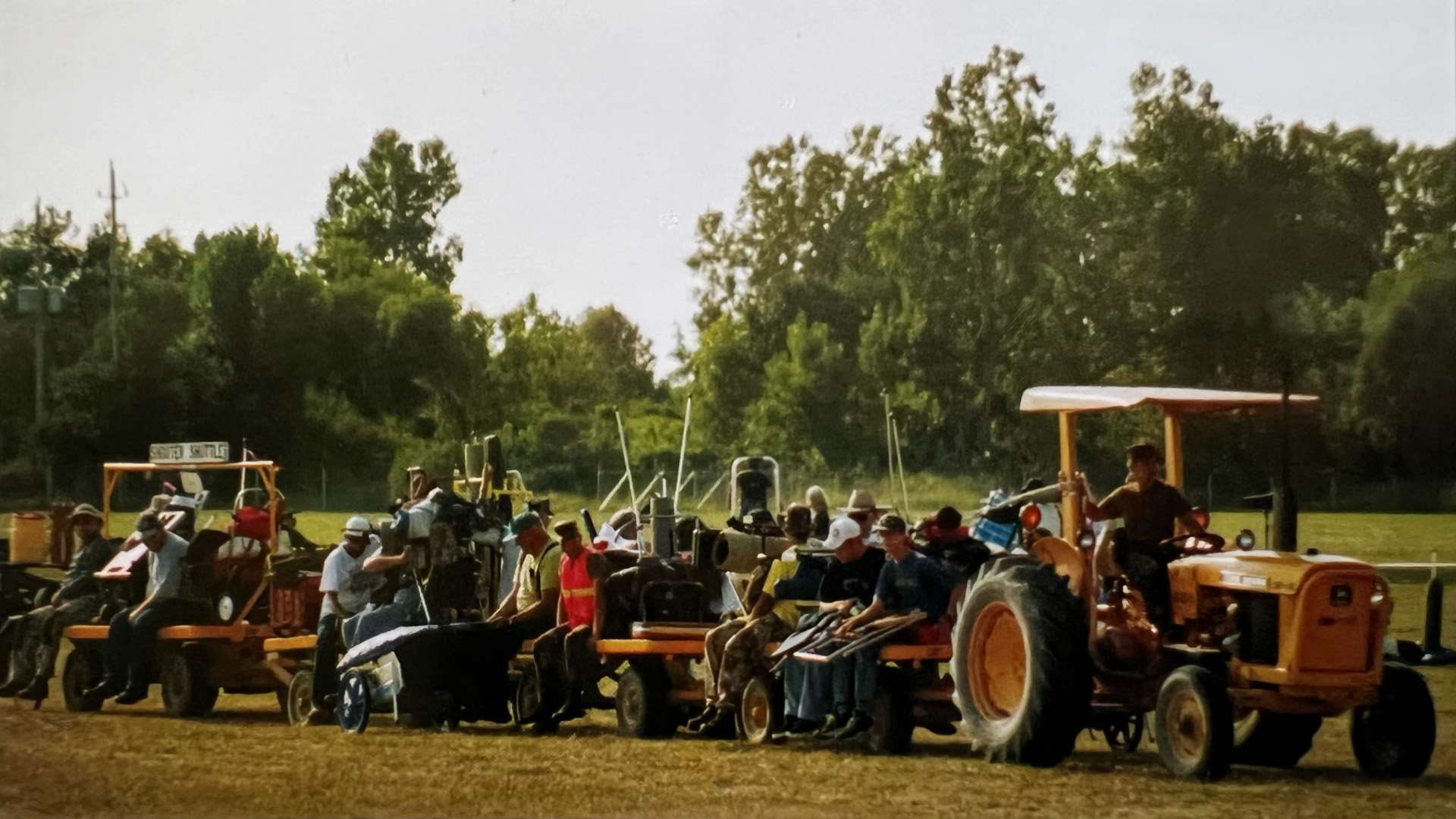 Competitors and gear carts ride a string of flatbed trailers towed by a yellow tractor across a grass field at Camp Perry