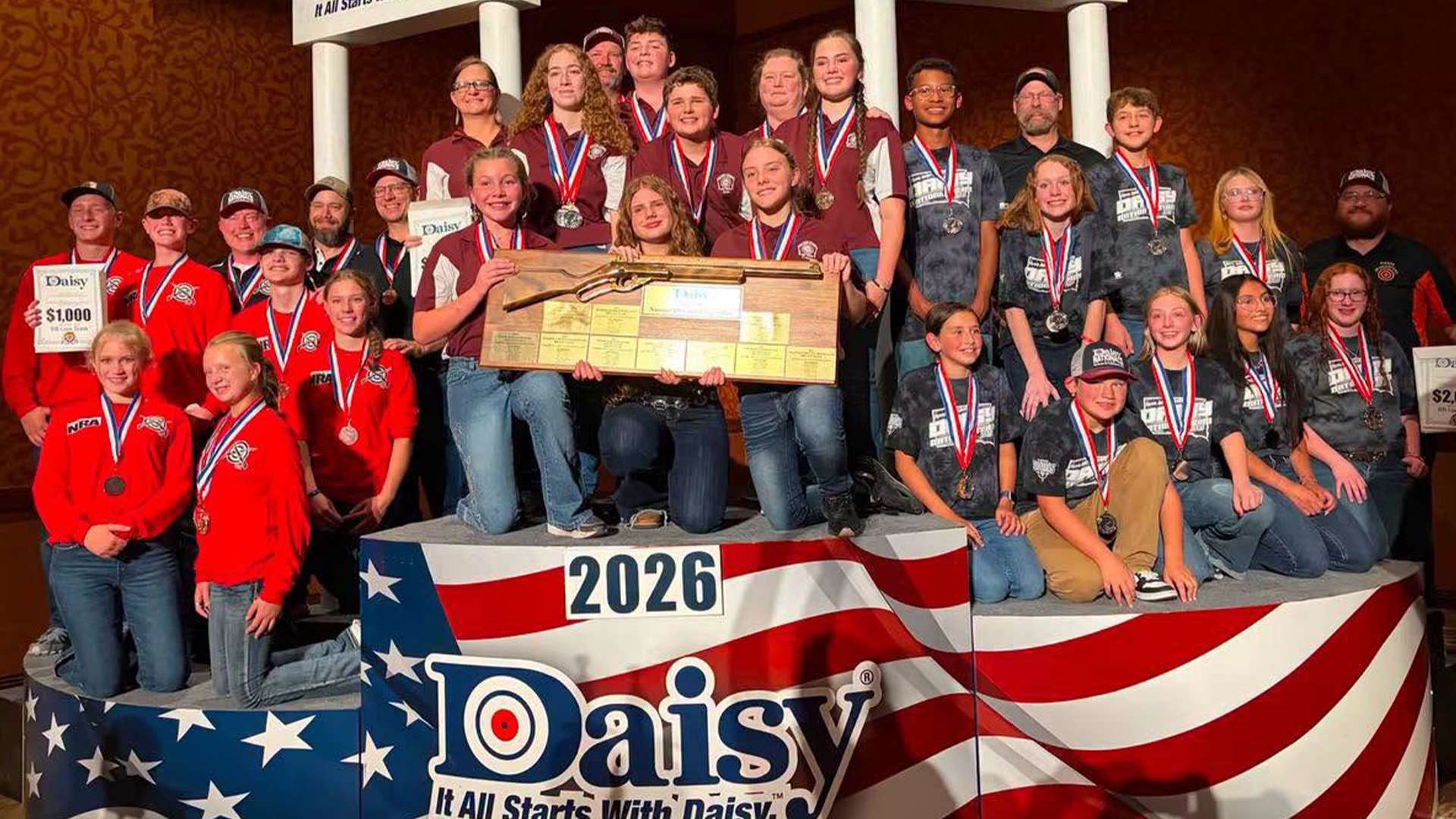 Three youth BB gun teams and their coaches stand and kneel together on a large red, white and blue podium at the 2026 Daisy National BB Gun Championship Match in Rogers, Arkansas