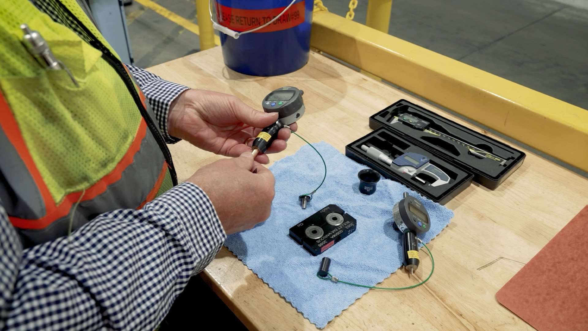 Winchester quality technician in a safety vest using a digital dial indicator to measure a bullet, with micrometers, calipers and go/no-go bullet diameter gauges on a workbench