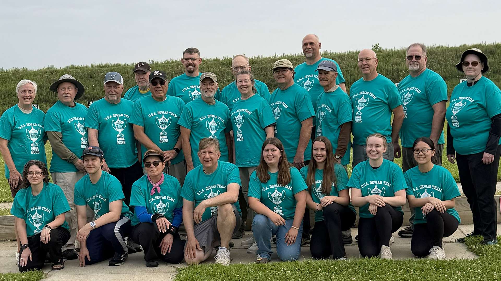 Roughly two dozen competitors in teal U.S.A. Dewar Team shirts pose in three rows on the firing line at the Cardinal Shooting Center