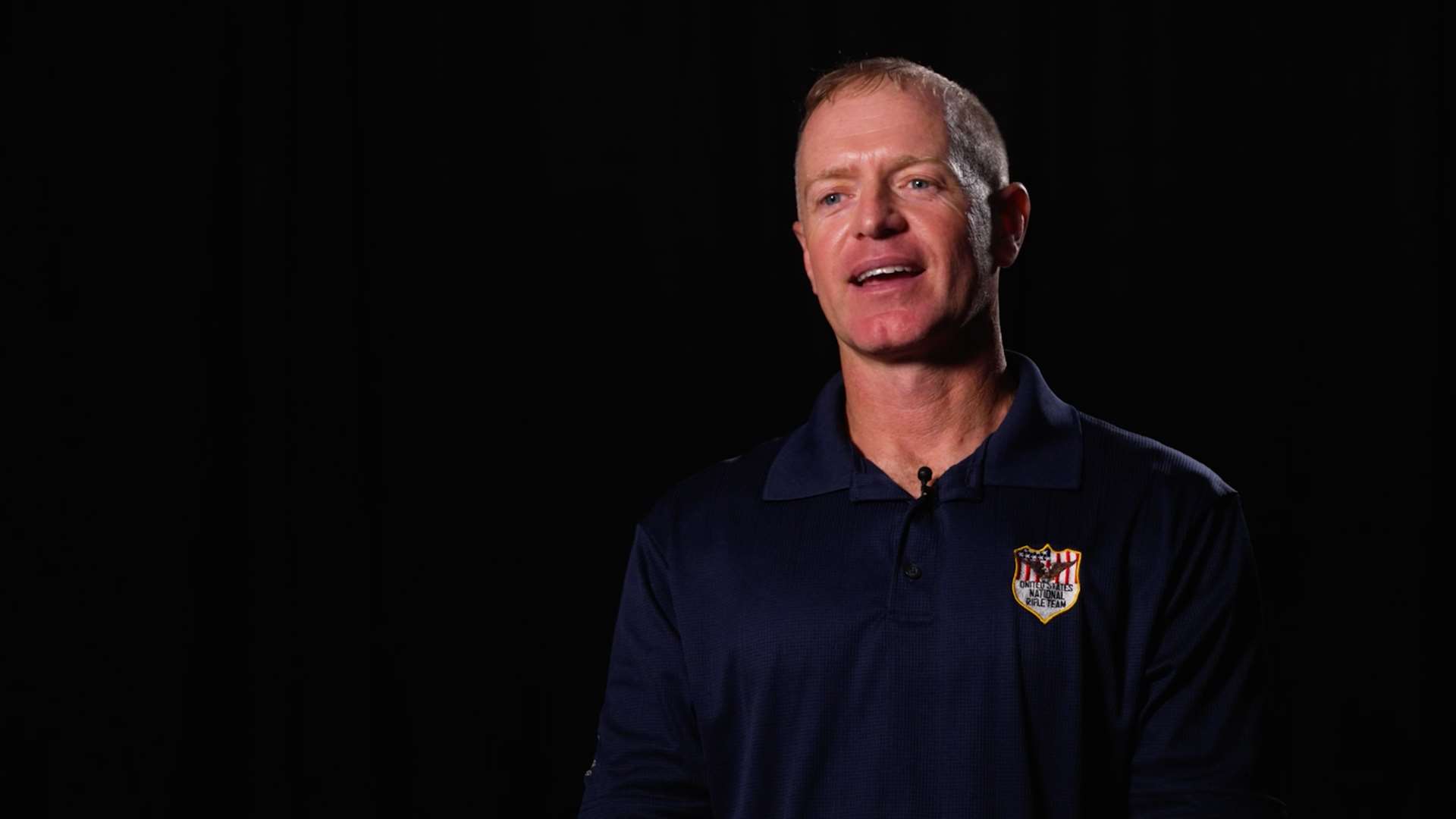 Brandon Green, wearing a navy U.S. National Rifle Team polo shirt, speaks to an interviewer against a black studio backdrop