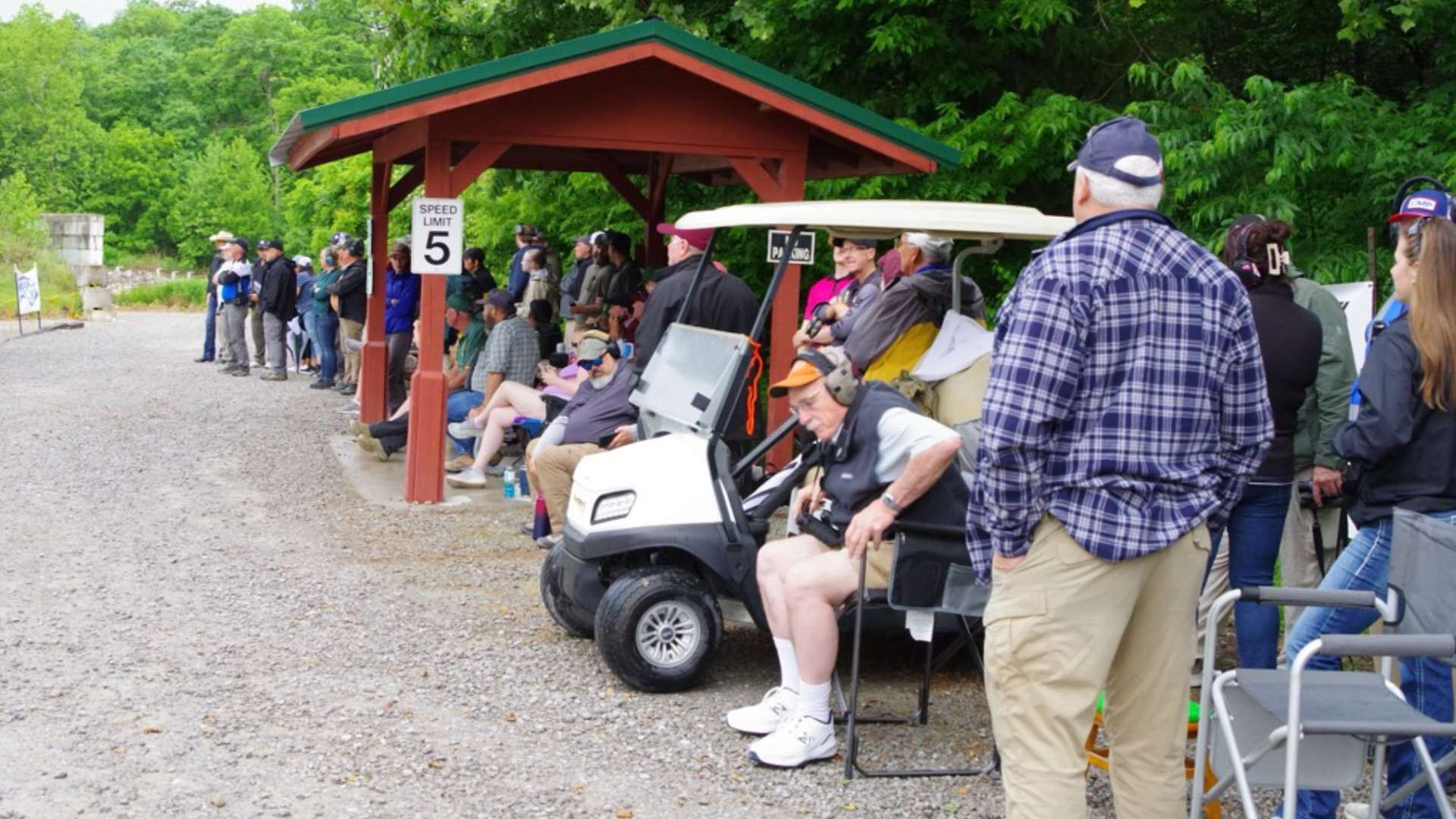 Competitors and match staff stand and sit along a gravel range road beside a covered shelter and a parked golf cart at the 2026 CMP Bianchi Cup at Green Valley Rifle and Pistol Club, with green trees in the background