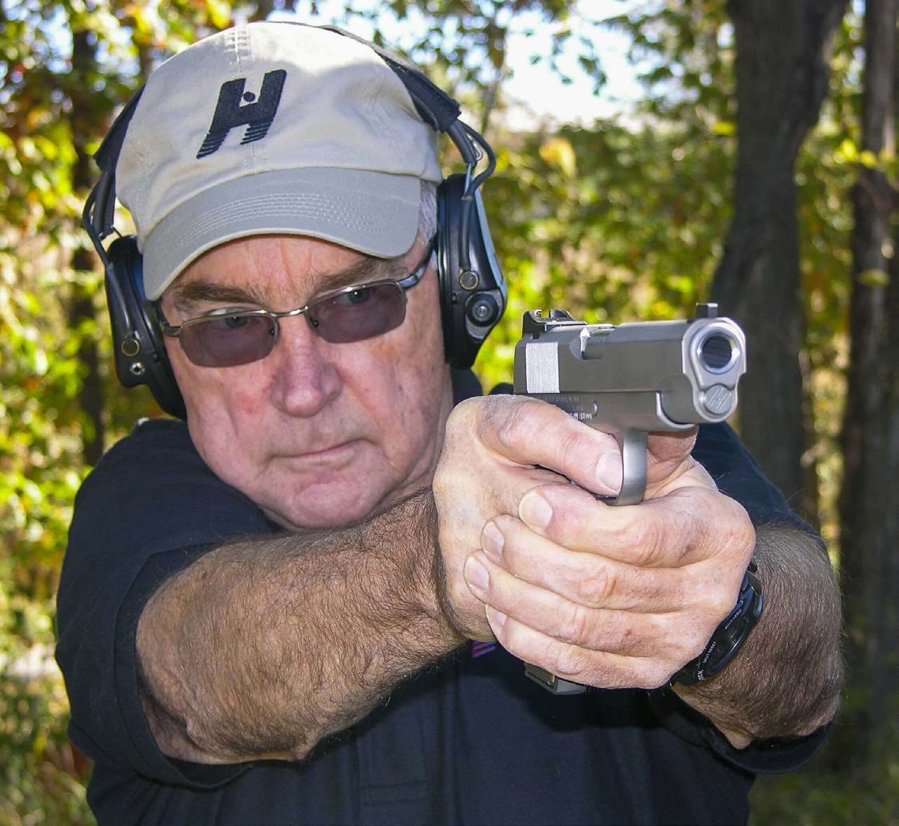 Gunsmith Richard Heinie firing a stainless 1911 pistol at an outdoor range, wearing a tan Heinie logo cap, sunglasses and earmuffs