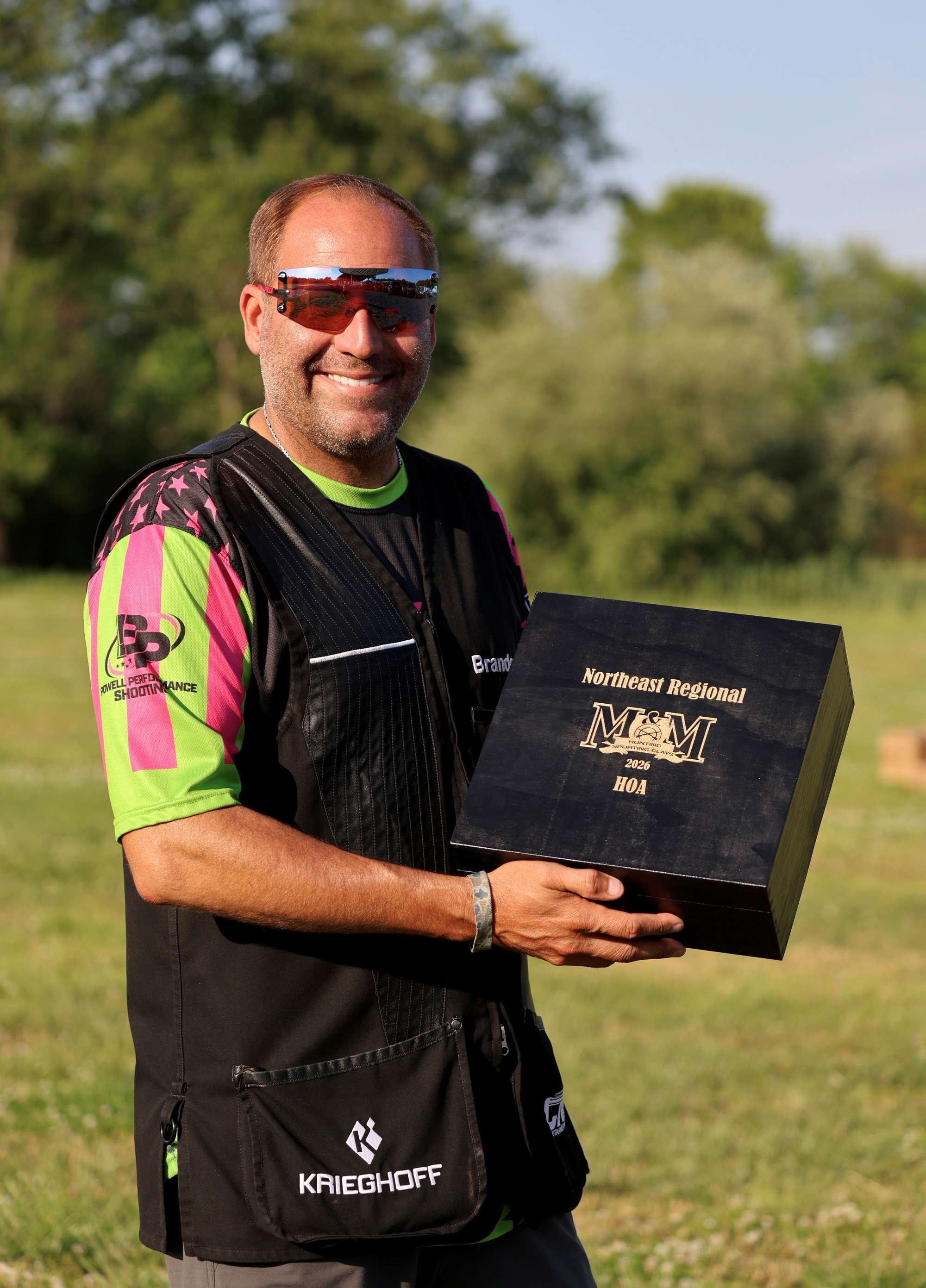 Brandon Powell smiles while holding the black 2026 M&amp;M Northeast Regional HOA trophy box, wearing a pink and green Powell Performance Shooting jersey under a black shooting vest