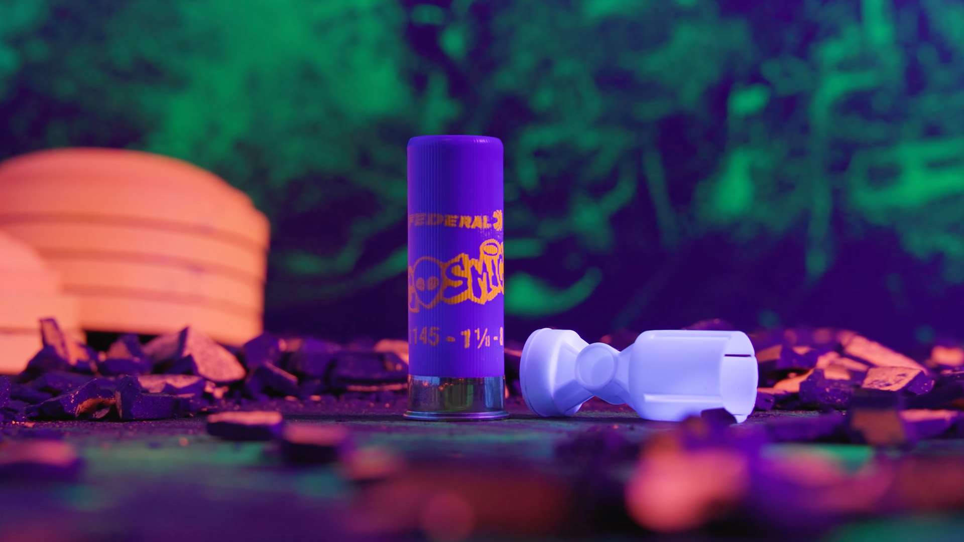 Federal Cosmic shotshell standing upright next to a white Podium wad under UV light, with a stack of clay targets glowing orange behind