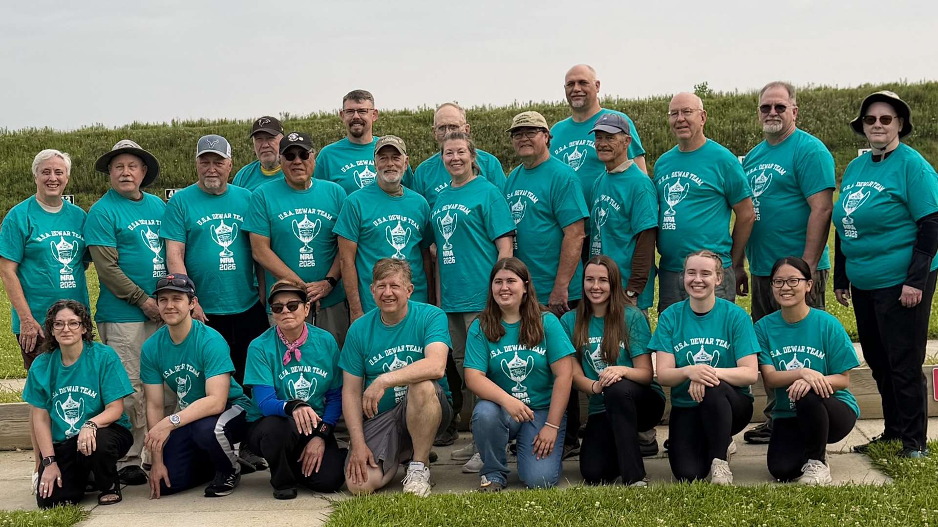 Roughly two dozen smallbore competitors in teal U.S.A. Dewar Team shirts pose in three rows on the firing line at the Cardinal Shooting Center, with a grass berm and target frames behind them