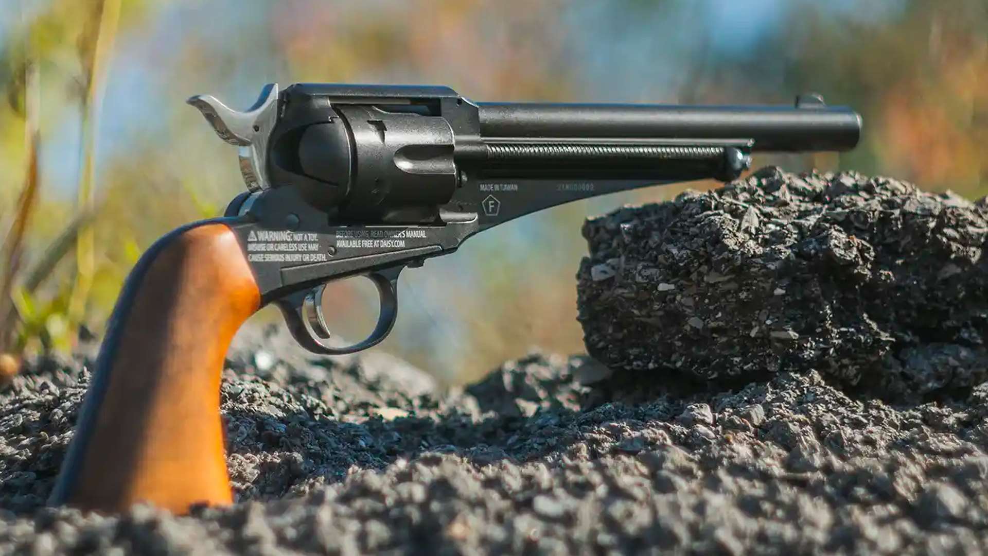 Winchester Single Action Western Revolver resting on rocks outdoors with a blurred autumn background showing its rifled barrel and faux wood grip