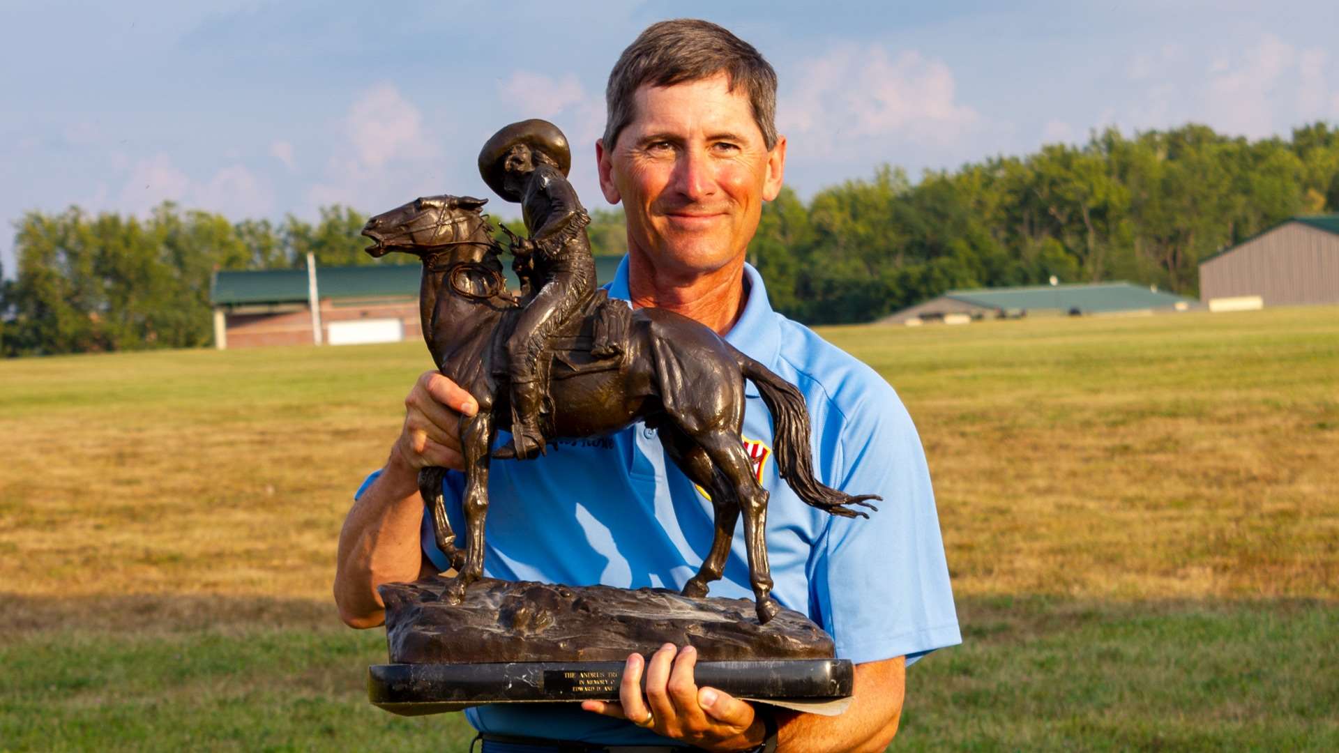 Charles Rowe holds the Edward D. Andrus Memorial Trophy, a bronze statue of a mounted scout on a horse, on a grass range at Camp Atterbury
