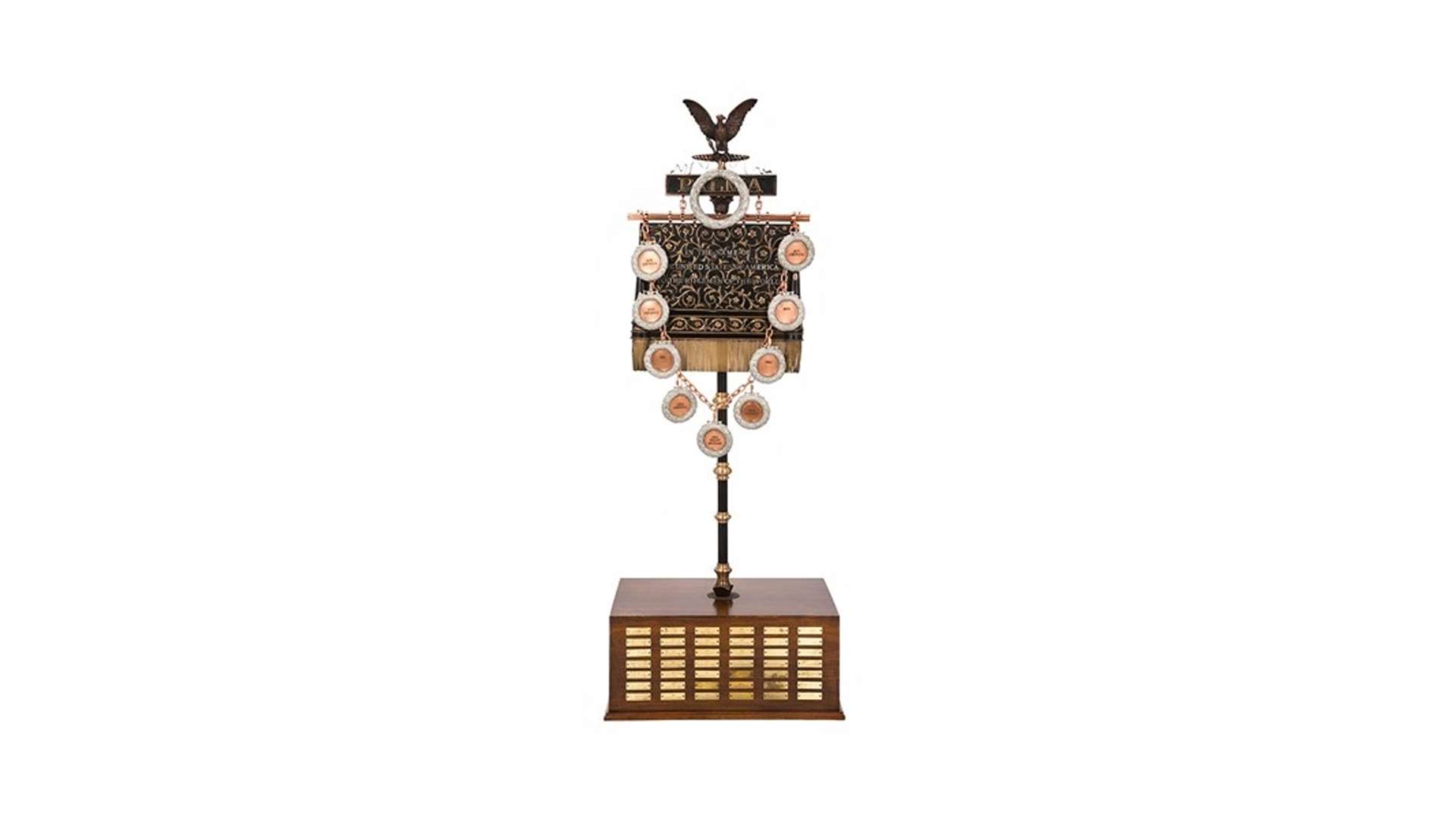 Full-length photograph of the current Palma Trophy standing on a wooden base with engraved winner plaques, topped by a bronze eagle and hung with circular medallions