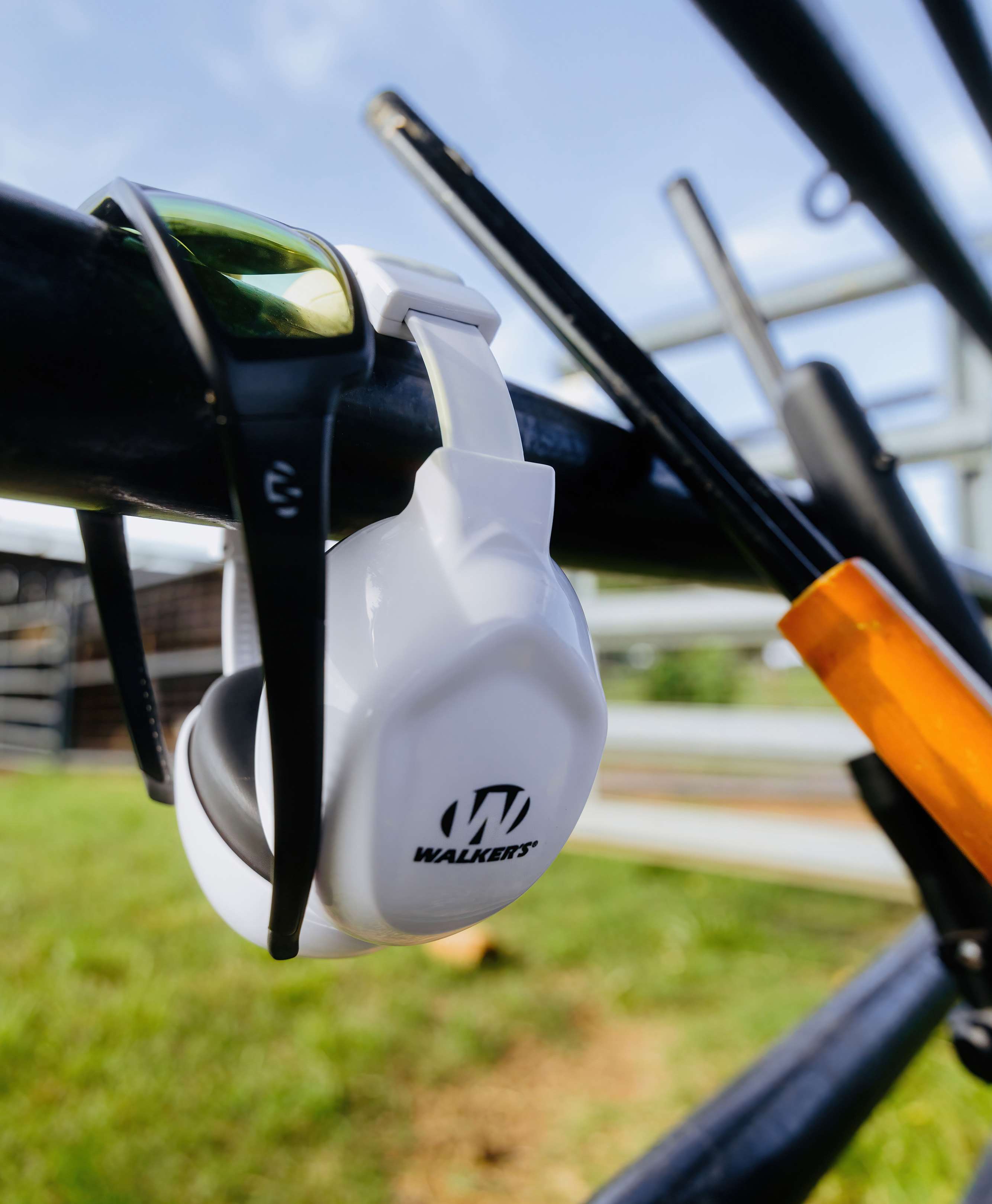 Walker's Boomies Passive Muffs in white, hanging by the headband from a black frame outdoors, showing the contoured low-profile ear cup with Walker's logo and padded cushion