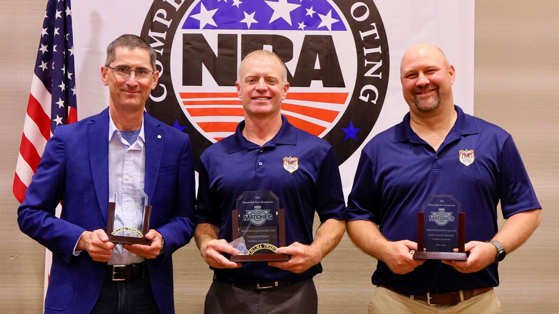 Charles Rowe, Brandon Green and Joe Bakies hold their Palma Individual Match awards at the 2026 NRA Long Range Awards ceremony with an NRA banner behind them