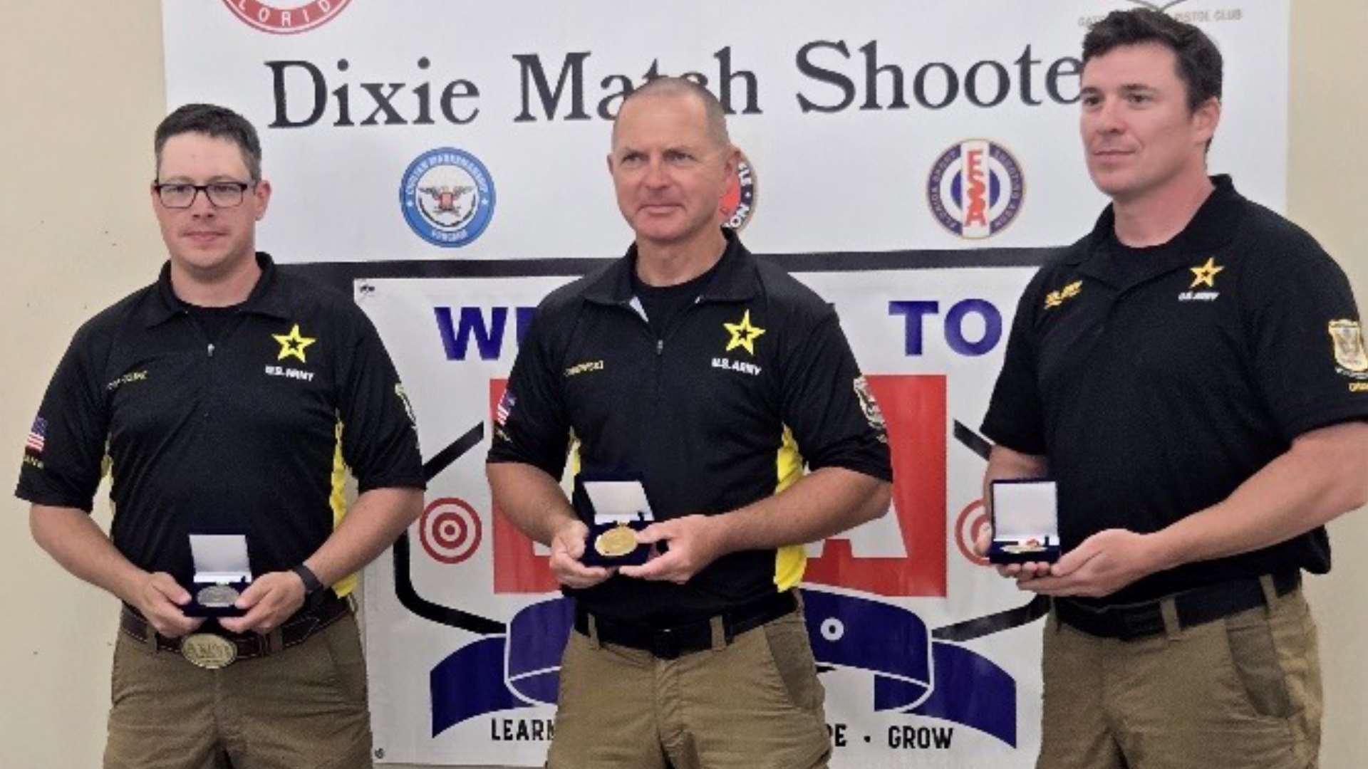 Three U.S. Army Marksmanship Unit shooters in matching black polos hold open medal cases in front of the Dixie Match Shooters banner, with Greg Markowski center holding the gold