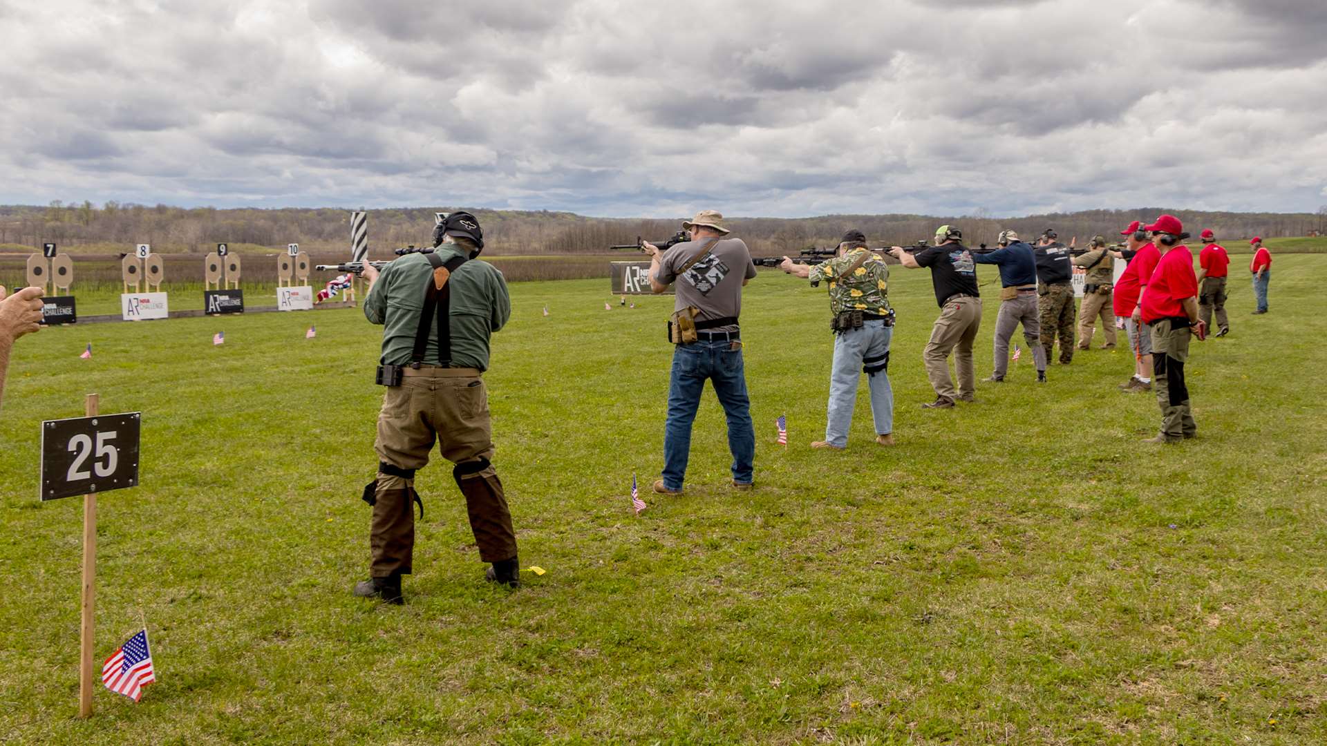 Line of competitors shooting AR-15s from standing position at NRA America's Rifle Challenge cardboard targets during ARC Level 1 stage with American flags marking the firing line