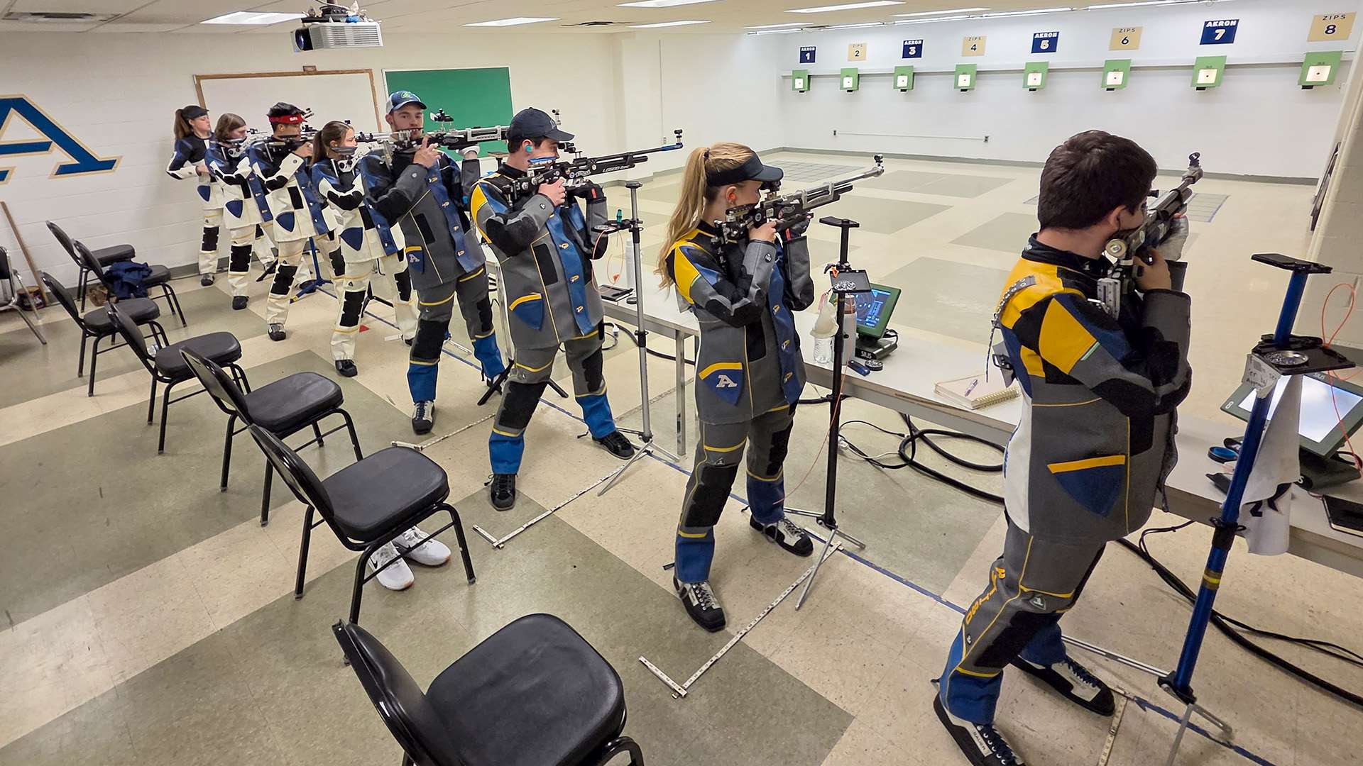 A line of collegiate rifle athletes aim air rifles from the standing position on an indoor range, Navy in white-and-navy jackets in the background and Akron in gold, blue and gray in the foreground