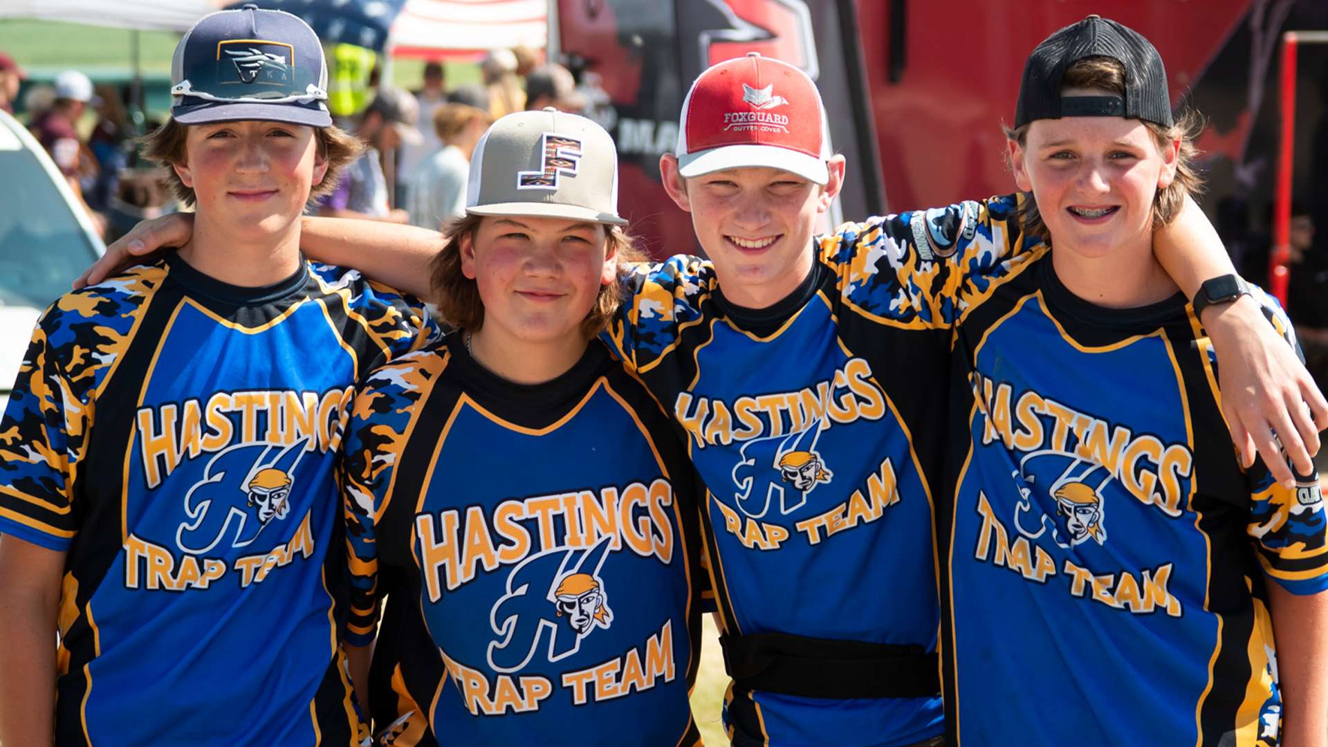 Four young shooters in blue and gold Hastings Trap Team jerseys and ball caps stand arm in arm, smiling, at an outdoor shooting event