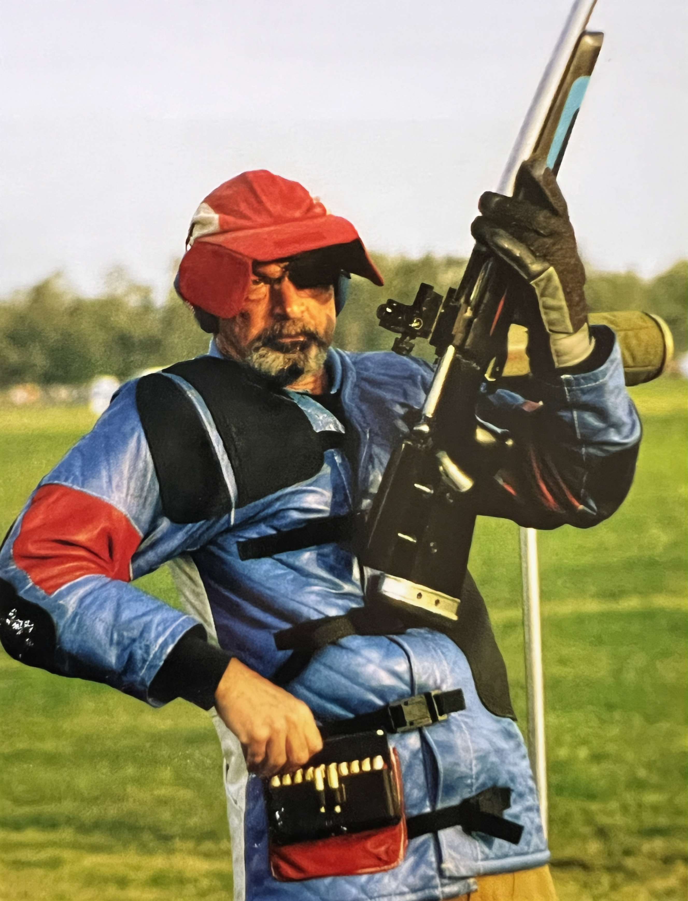 Tom Whitaker, bearded and wearing a red cap with an eye patch and a blue shooting coat, steadies a match rifle upright with a cartridge belt at his waist