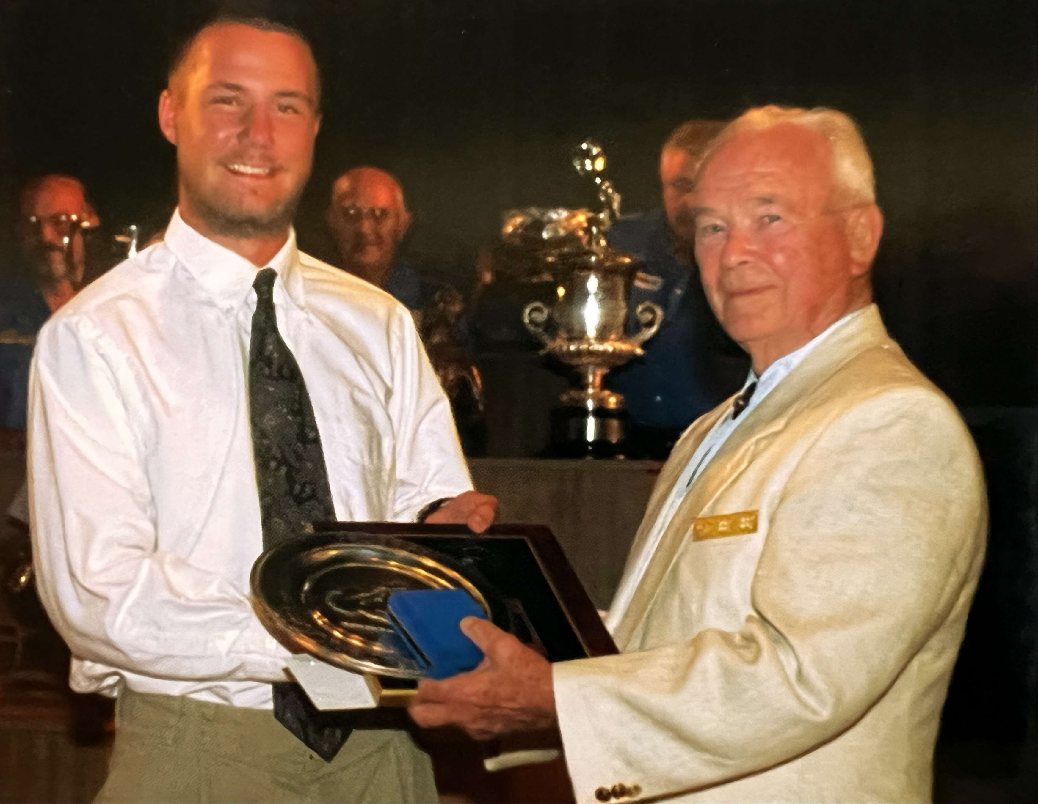Col. Walter Walsh, in a cream blazer, presents a silver tray and plaque to John Bickar in a white shirt and tie, a large trophy visible behind them