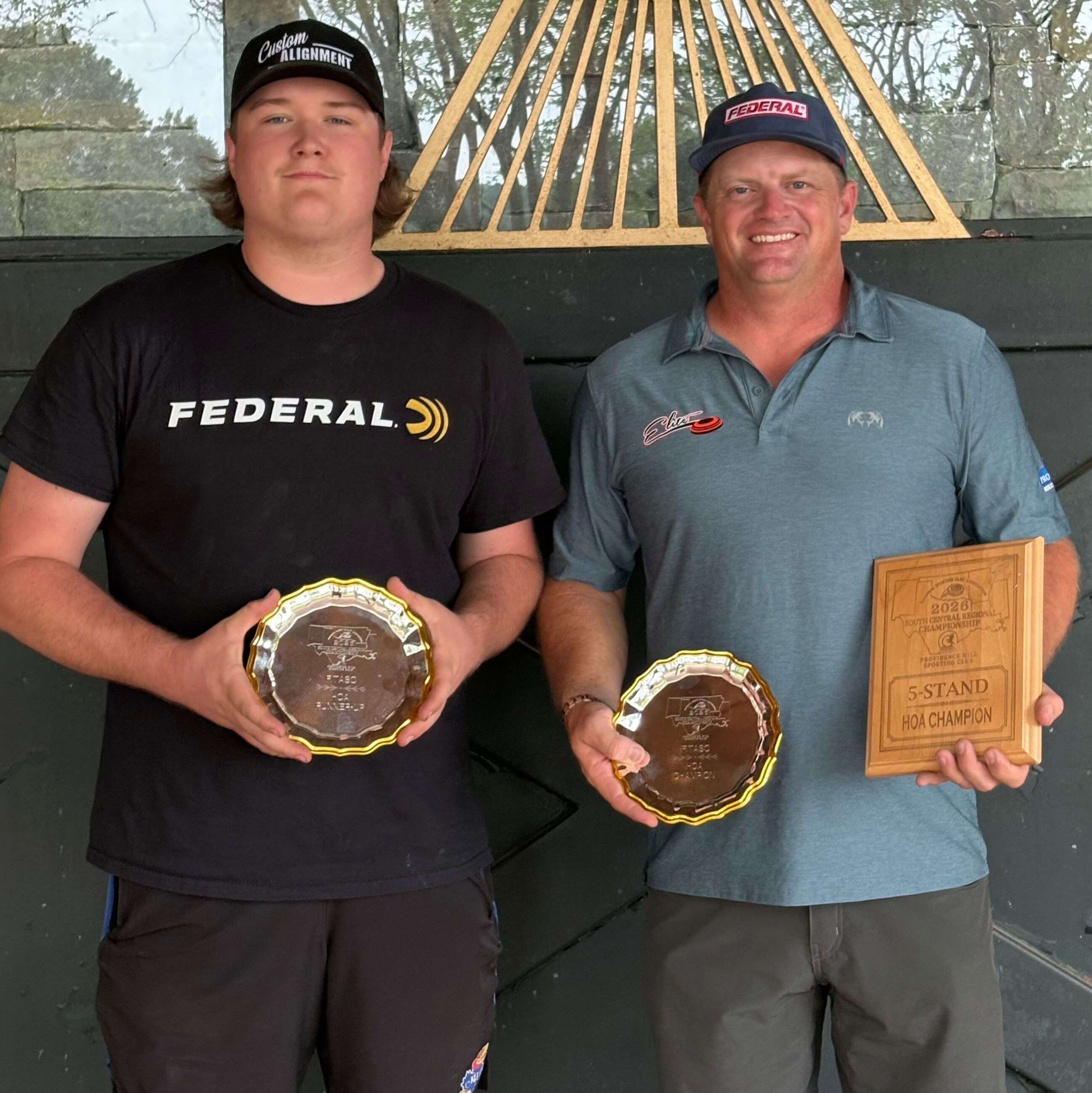 Two shotgun shooters stand against a dark wall; one holds a silver plate, the other holds a silver plate and a wooden plaque reading "5-Stand HOA Champion."