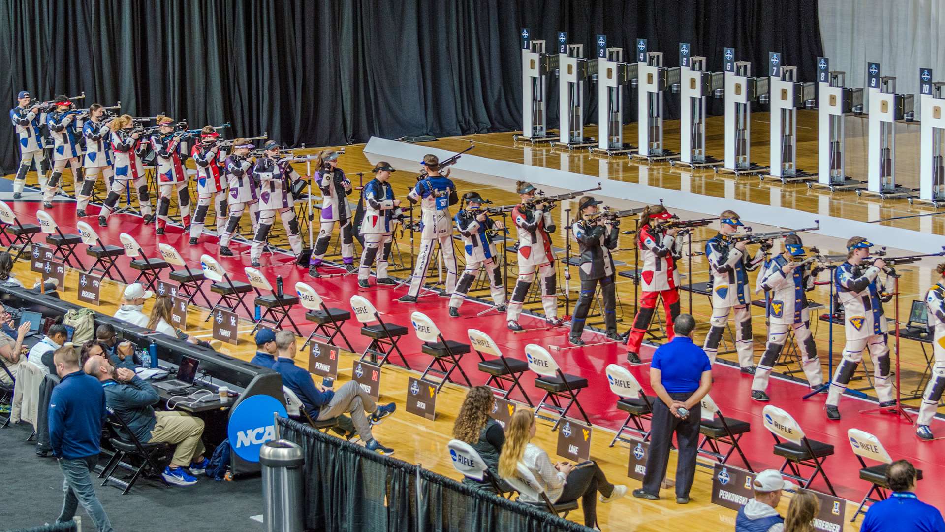 A long line of college rifle shooters in shooting jackets aims air rifles from the standing position at the 2026 NCAA Rifle Championship