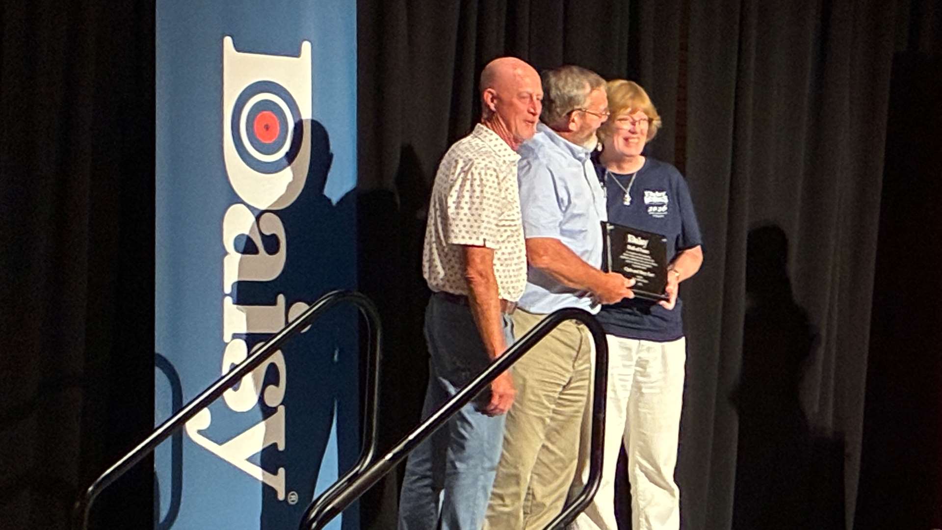 Side view of Clyde and Mary Furr holding their Hall of Fame plaque on stage with Daisy CEO Keith Higginbotham during the induction ceremony at the 2026 Daisy Nationals