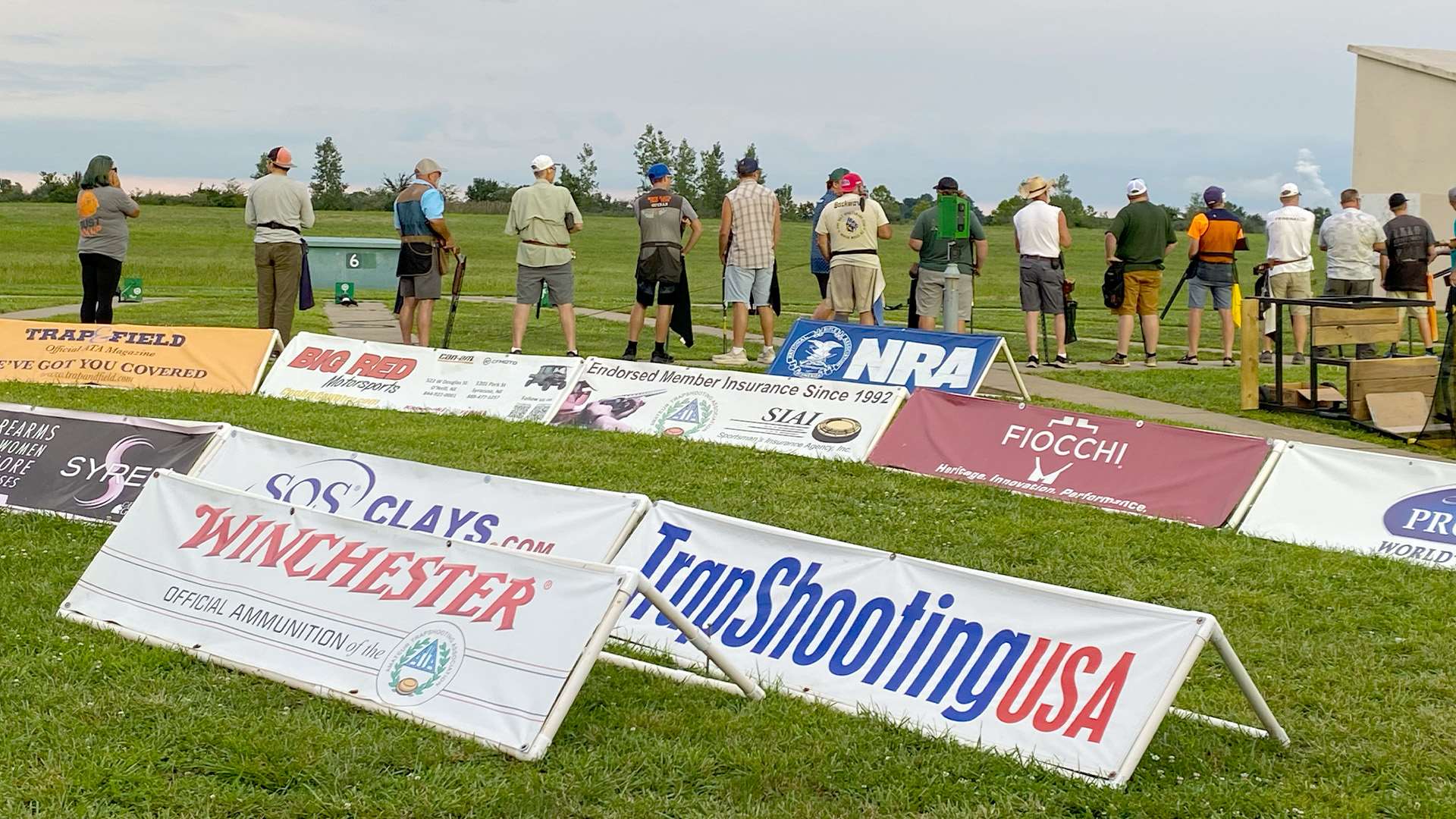 Sponsor banners for Winchester, the NRA, Fiocchi, TrapShooting USA and Trap &amp; Field line the grass in front of a full squad of trapshooters at the Grand American