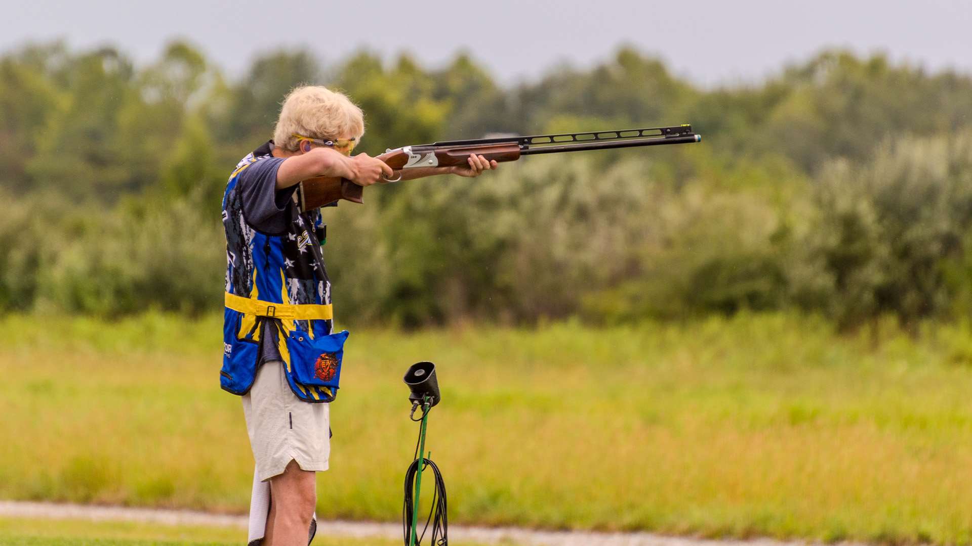 A trapshooter in a blue and yellow vest takes aim during the 2026 Grand American as light rain falls