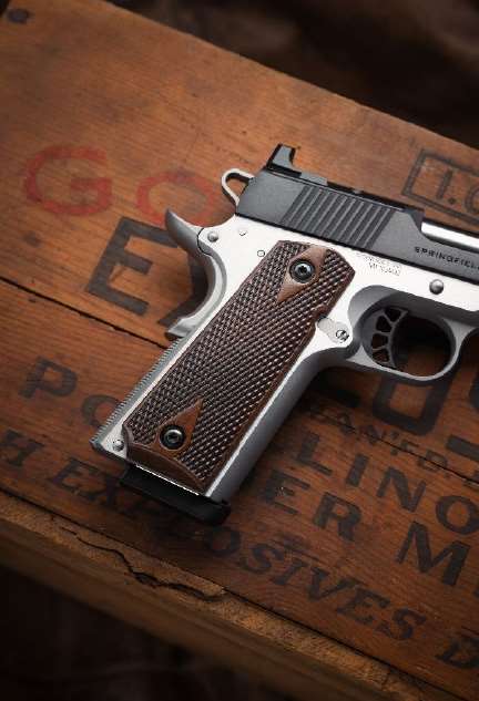 Close-up of a stainless 1911 grip frame fitted with checkered walnut double-diamond grip panels, resting on a weathered wooden crate with faded stenciled lettering