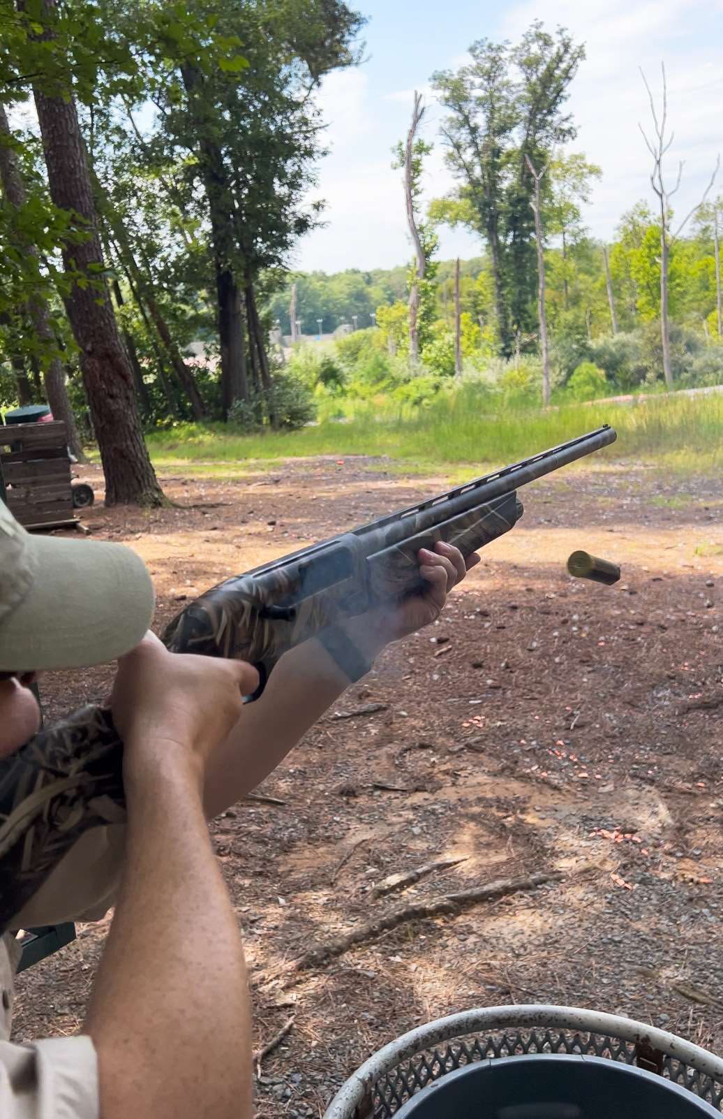 A shooter in a tan cap fires a camouflage TriStar Viper G3 semi-automatic shotgun on a wooded sporting clays course as an empty shotshell ejects in midair