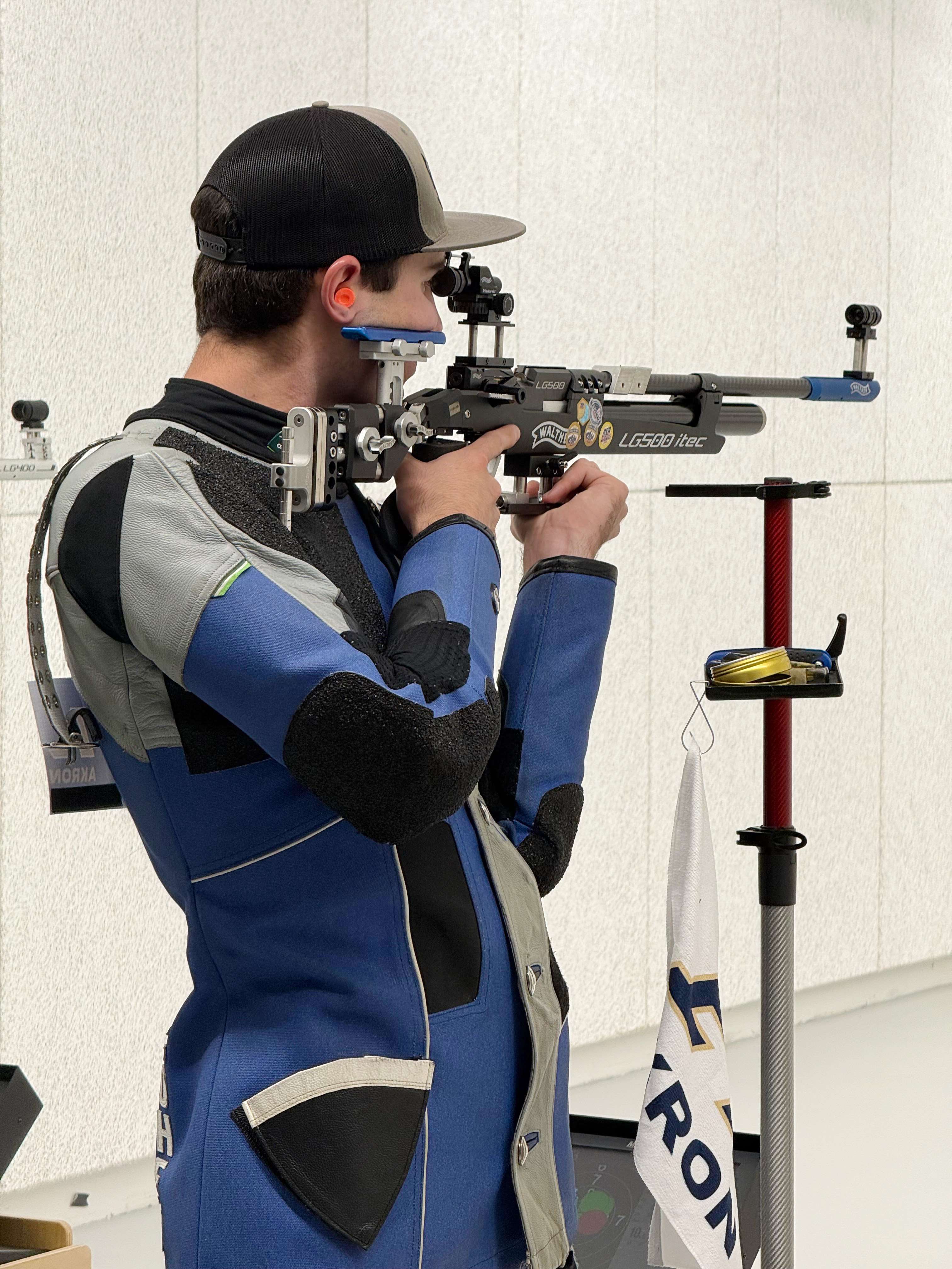 A male rifle athlete in a blue, gray and black shooting jacket aims a black Walther air rifle from the standing position beside a rifle stand draped with an Akron towel