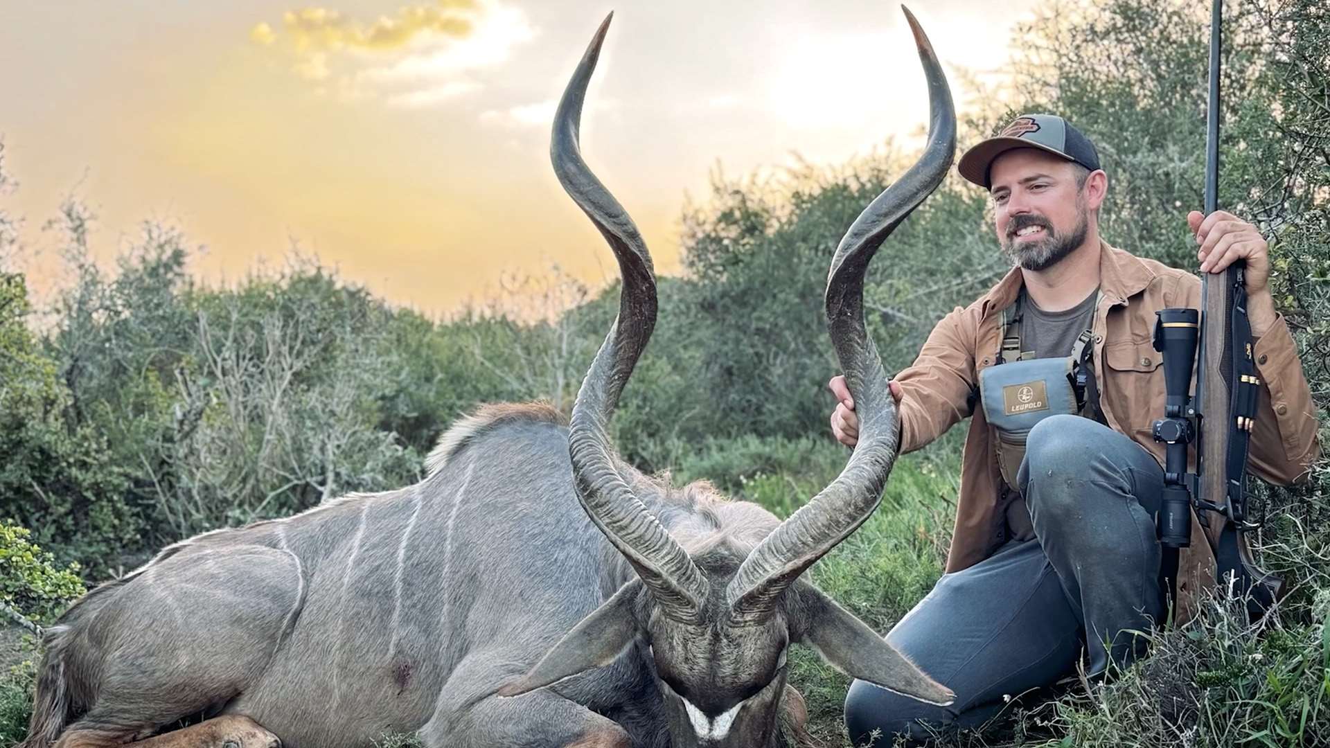 Hunter kneeling beside a kudu bull with long spiral horns, holding a scoped bolt-action rifle in African brush country at sunset