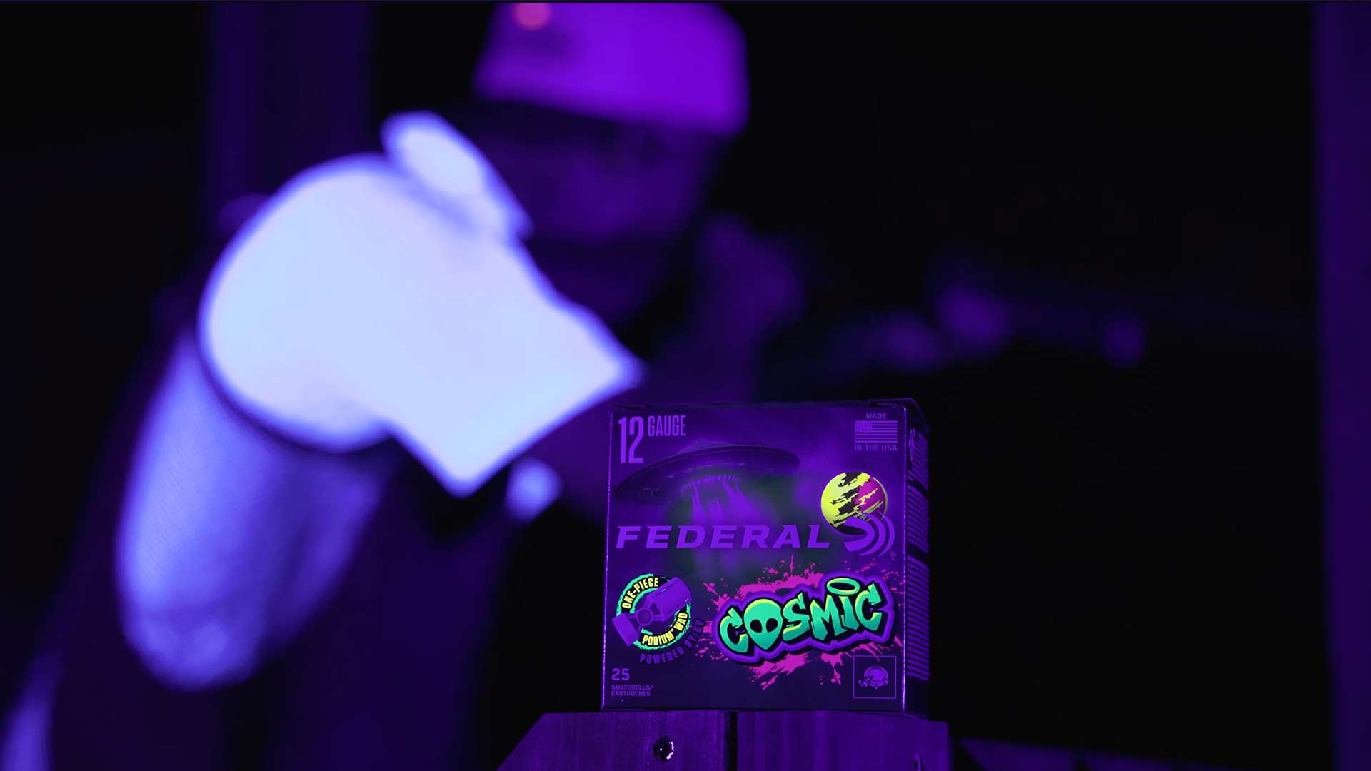 Federal Cosmic shotshell box on a post under purple lighting with a shooter and shotgun visible in the darkened background