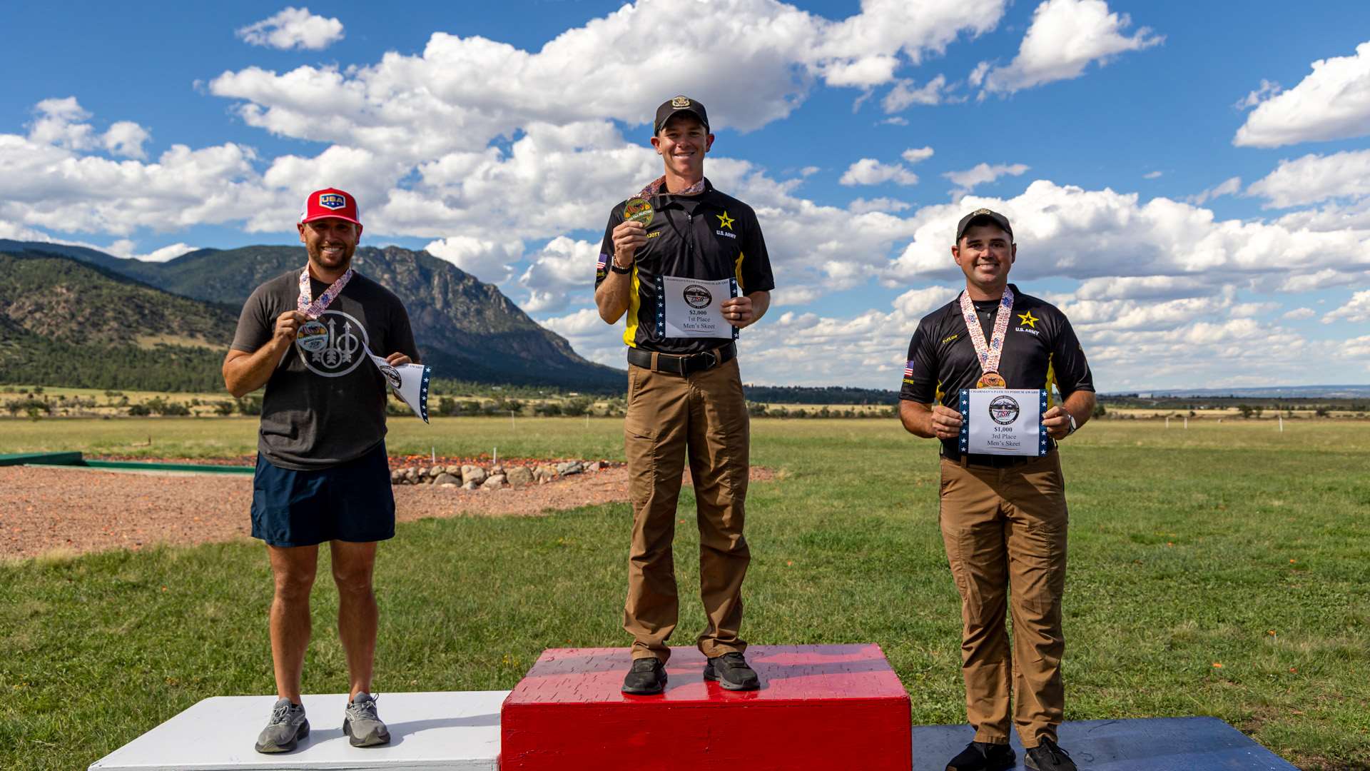Three male skeet shooters stand on a red, white and blue podium holding medals and award certificates, mountains behind them