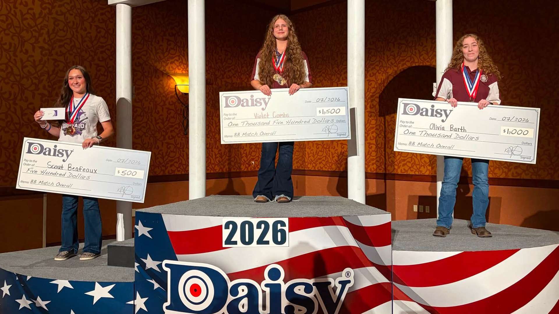 Three young female competitors stand on a red, white and blue podium holding oversized Daisy scholarship checks after the 2026 Daisy National BB Gun Championship Match in Rogers, Arkansas