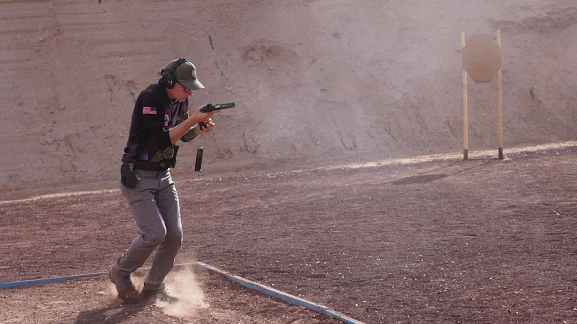 USPSA competitor reloads a pistol while moving through a dusty desert range, a spent magazine falling toward the ground beside a blue fault line