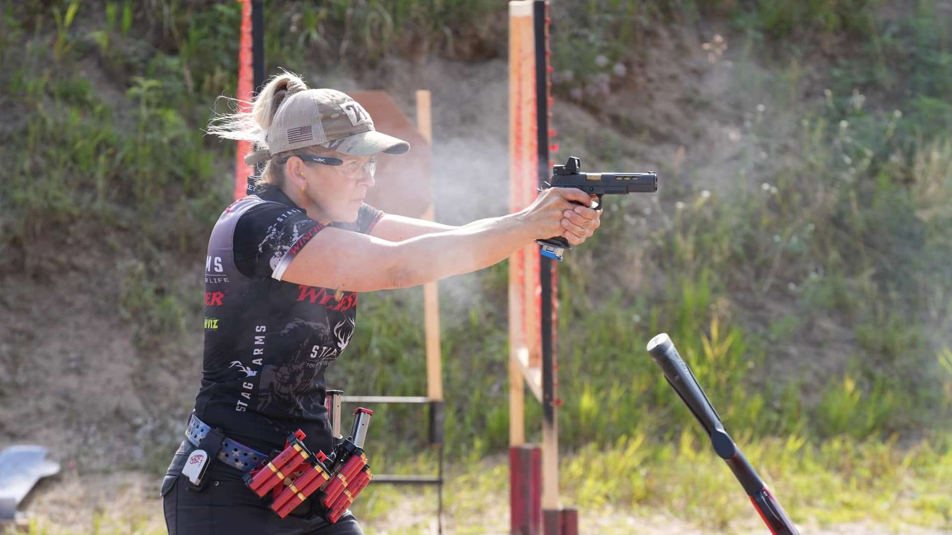 Woman in a competition jersey fires a pistol at a multigun match, shotshell caddies on her belt and a shotgun staged in the foreground