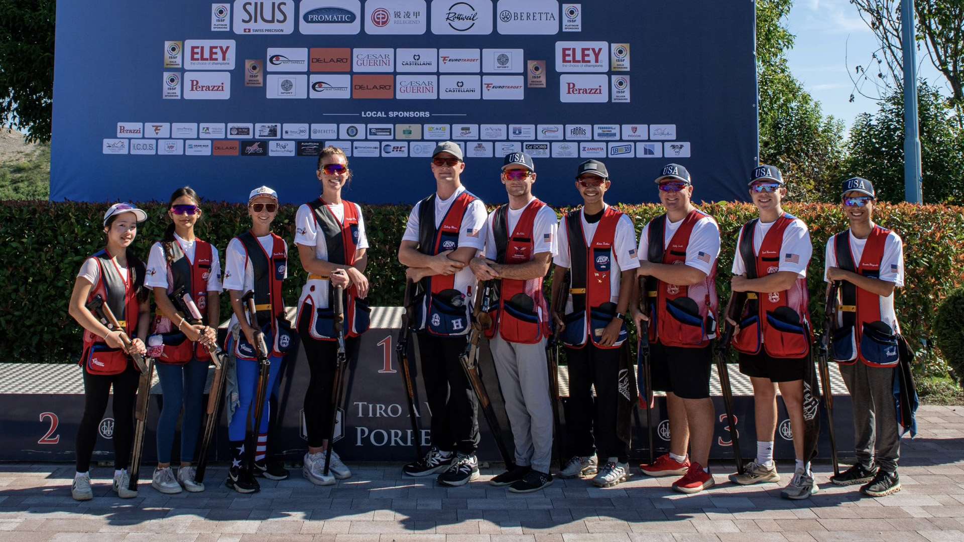 Ten U.S. junior shotgun athletes in red, white and blue shooting vests stand in front of the Porpetto podium with their shotguns, beneath a large sponsor backdrop