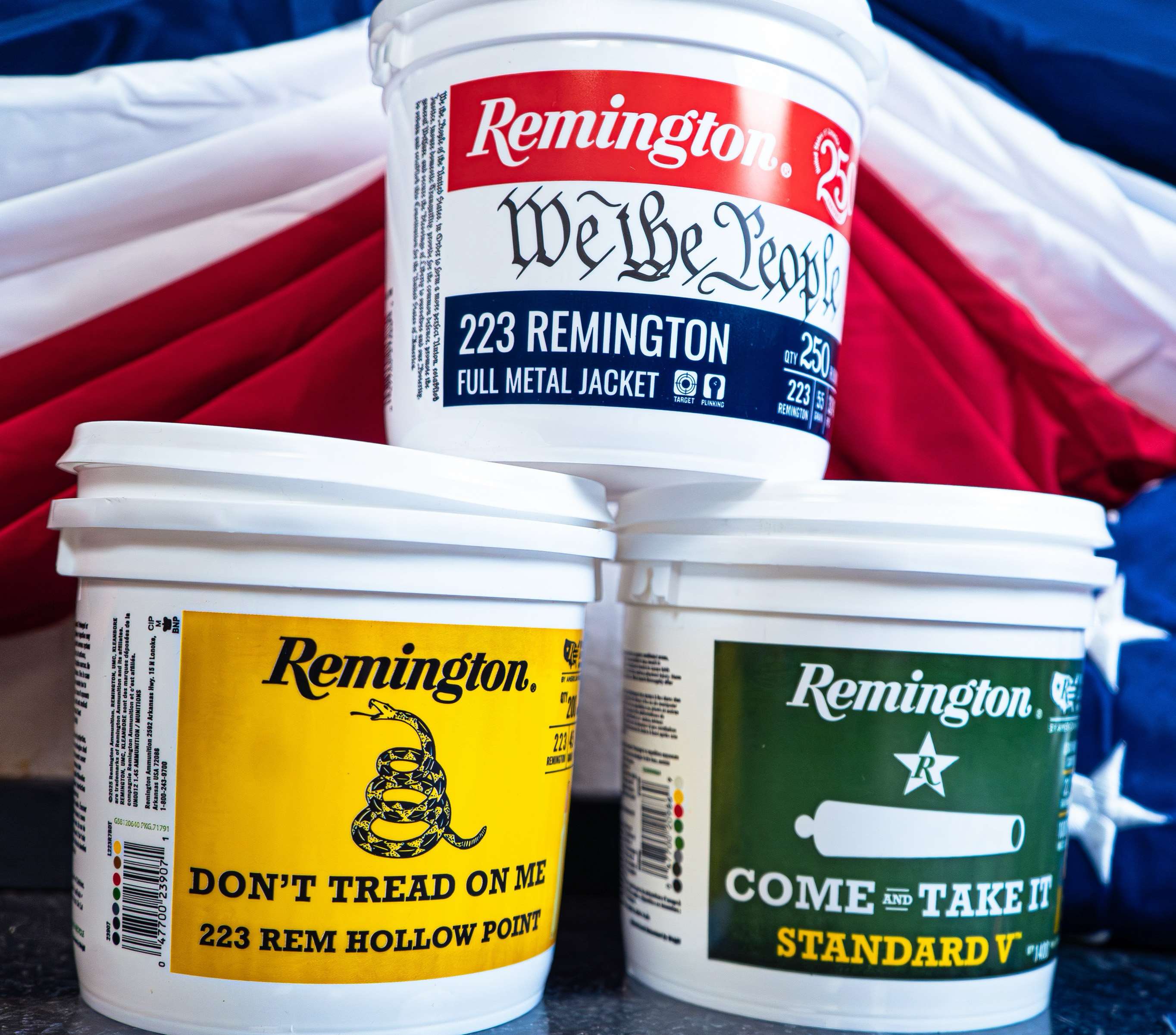 Three white Remington bulk ammunition buckets stacked in front of an American flag: a red, white and blue We the People .223 Remington bucket on top, with a yellow Don't Tread on Me bucket showing a coiled rattlesnake and a green Come and Take It bucket showing a white cannon and star below