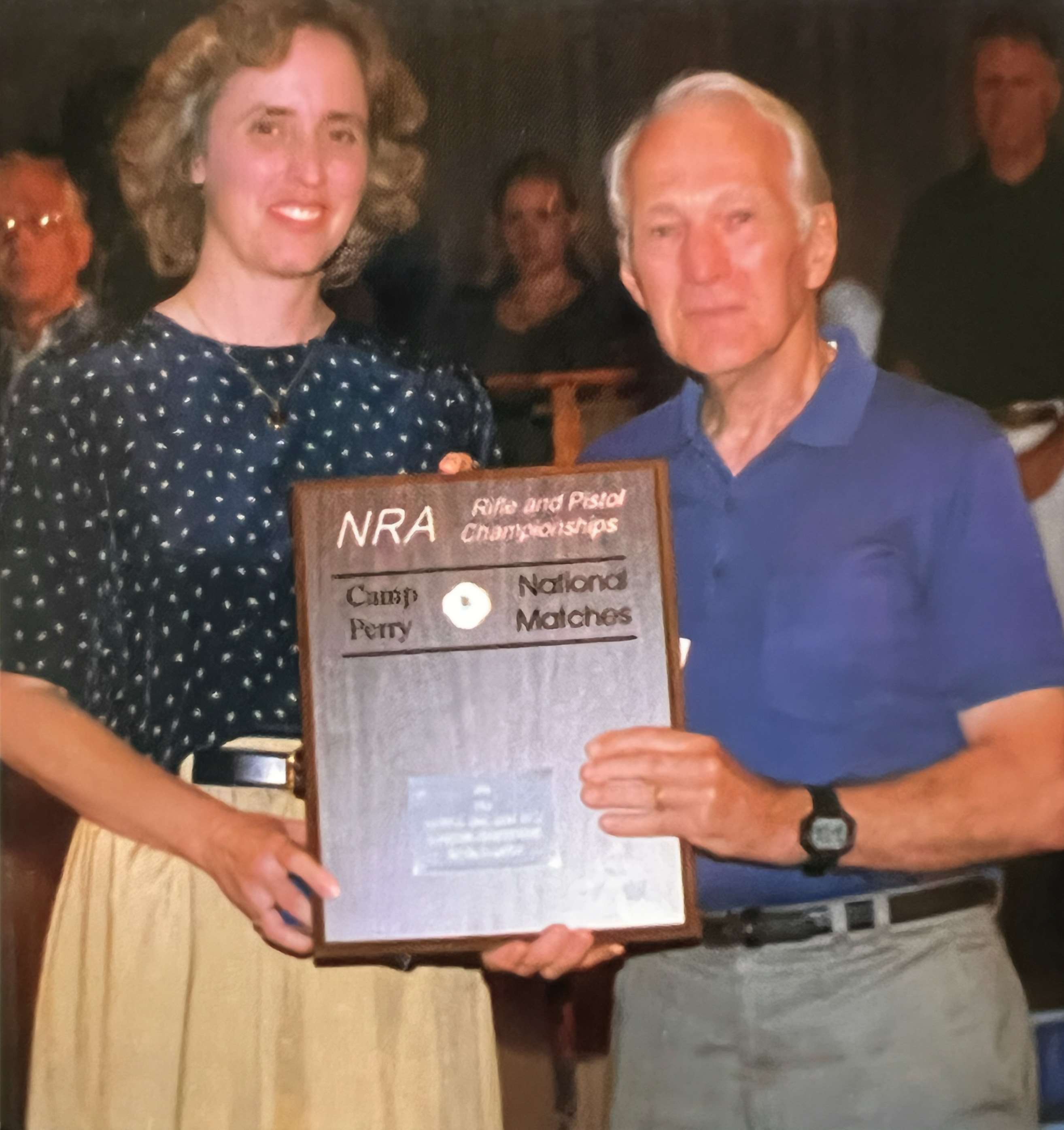 Edie Reynolds and Fred Cole hold a wooden NRA Rifle and Pistol Championships plaque between them at the Camp Perry awards ceremony