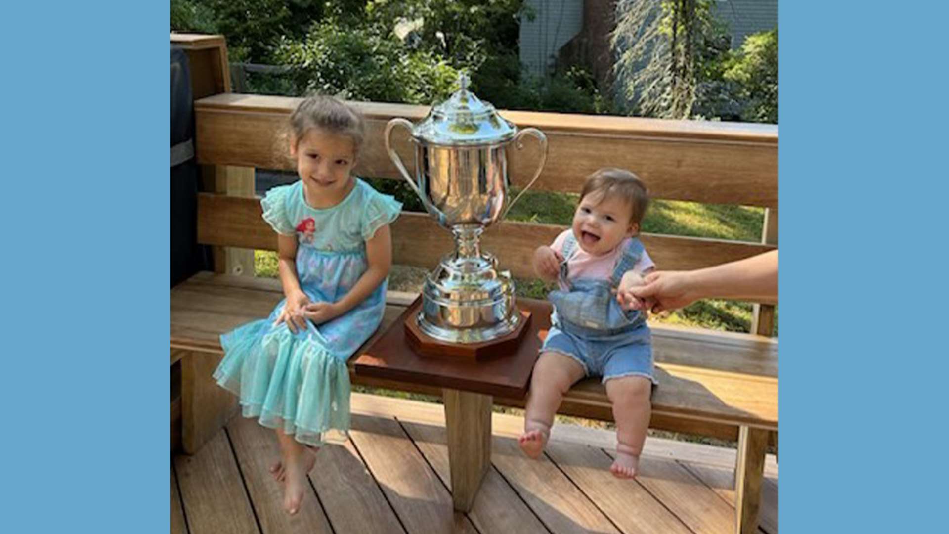 Two young children sit on either side of a large restored silver trophy cup mounted on a wooden base, resting on a wooden bench outdoors