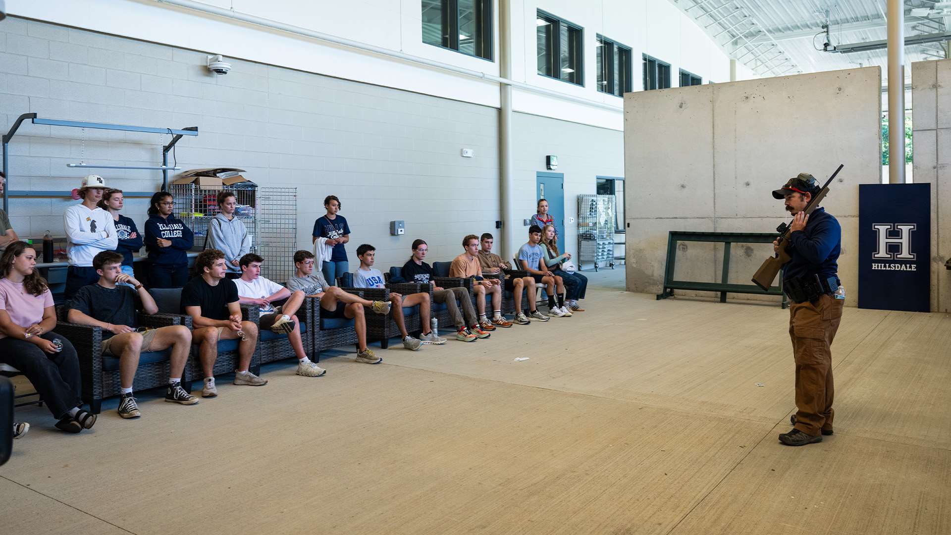 Instructor in a ball cap holding a scoped bolt-action rifle with its muzzle pointed upward speaks to college students seated and standing in front of a Hillsdale College banner