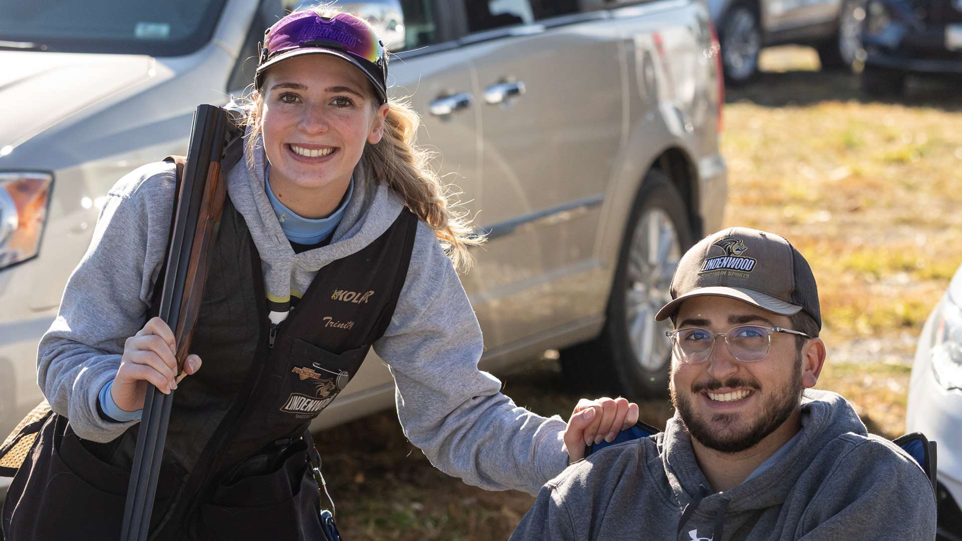 A smiling college shooter with a shotgun resting on her shoulder leans on the shoulder of a seated teammate in a parking area near the trap fields