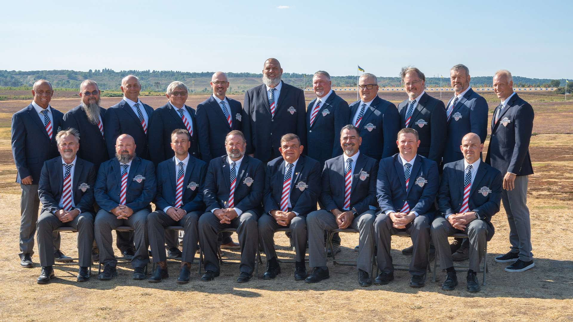 Formal portrait of the United States F-Class Open Rifle Team in navy blazers and striped ties, seated and standing in two rows on the range at Bisley with target butts visible behind them