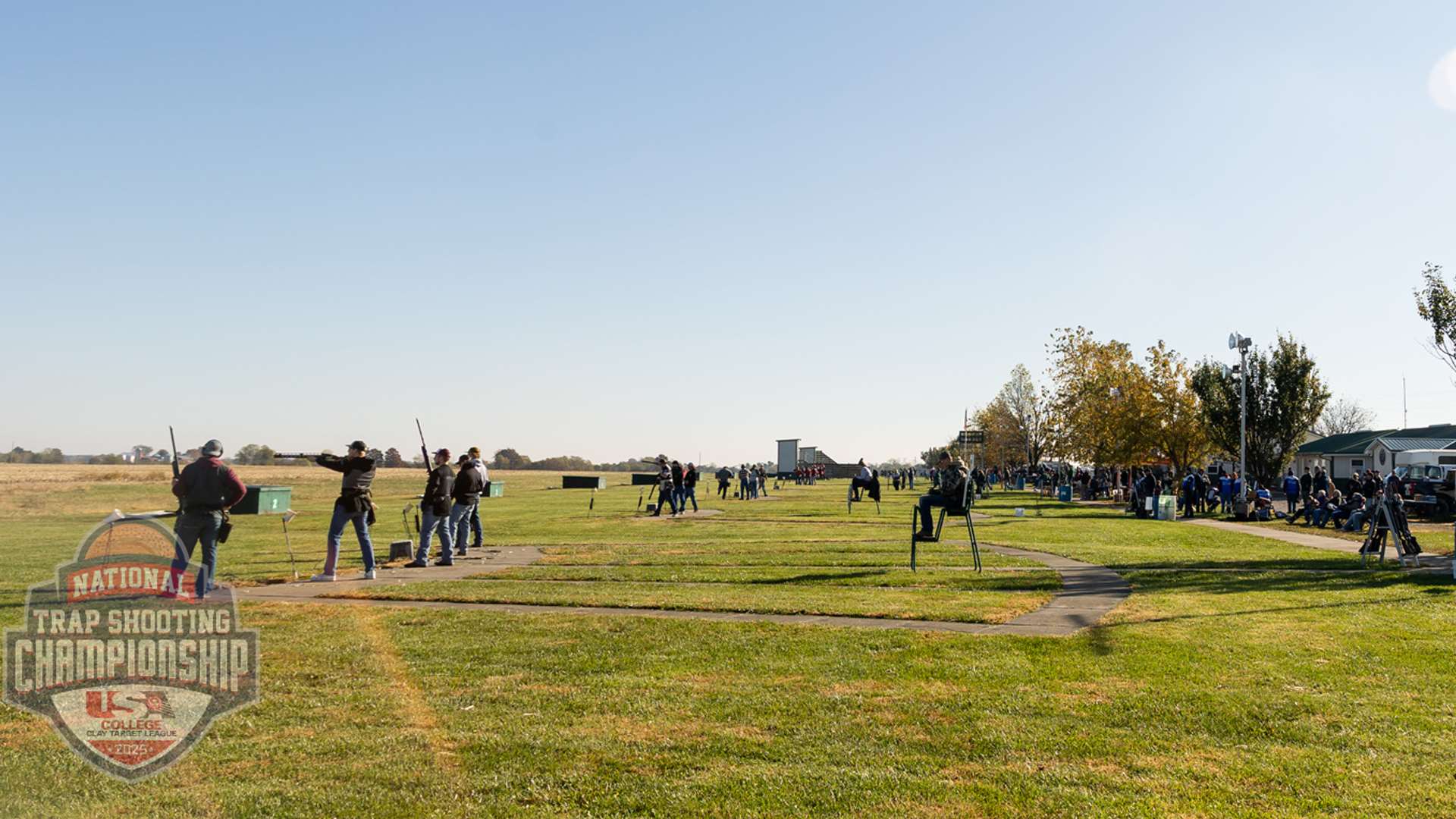 Rows of college athletes shoulder shotguns along a trap line on a sunny day with spectators and clubhouse buildings in the background