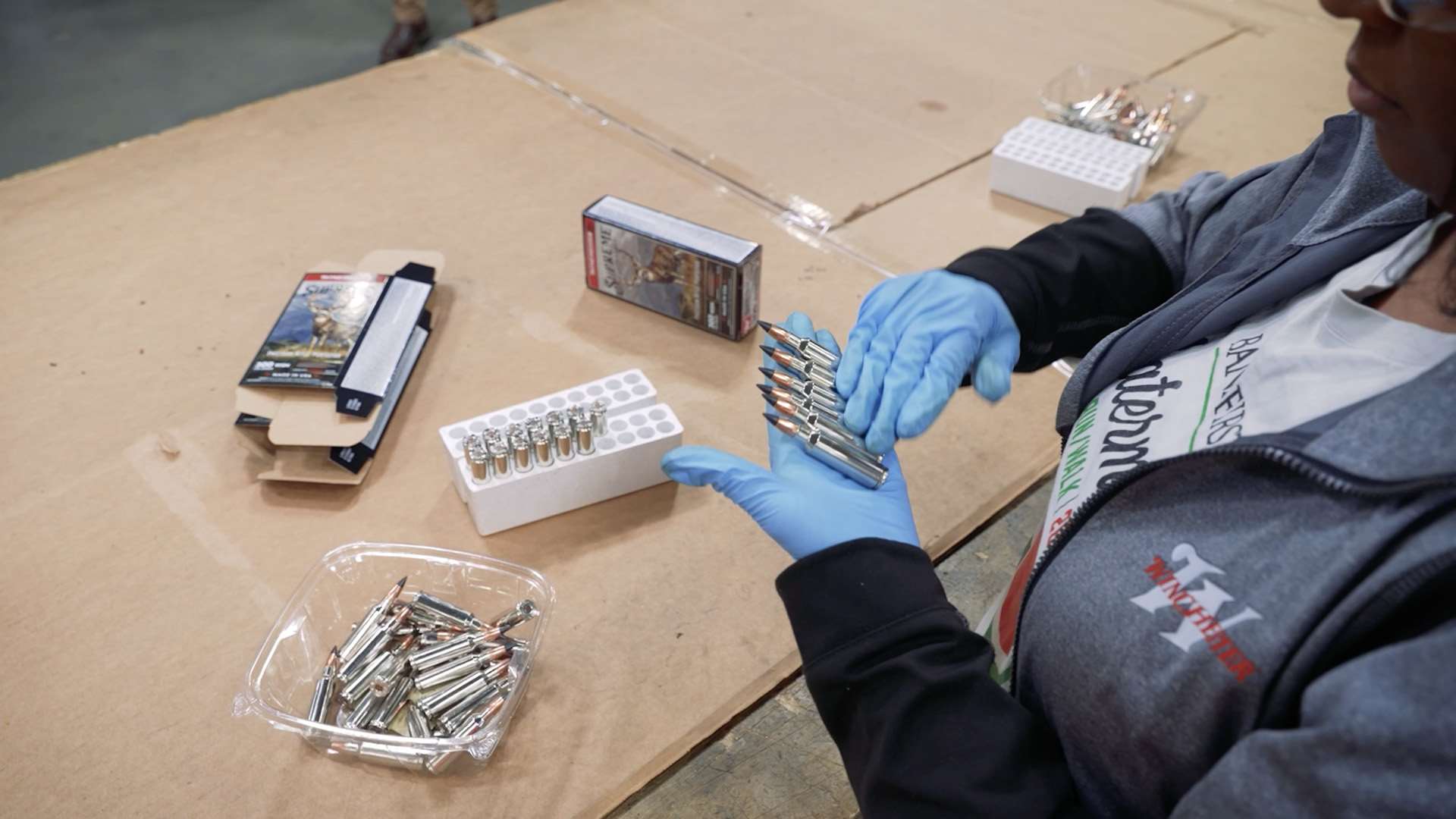 Winchester employee wearing blue gloves hand-inspecting and packing nickel-cased Supreme Long Range rifle cartridges into 20-round boxes at the factory