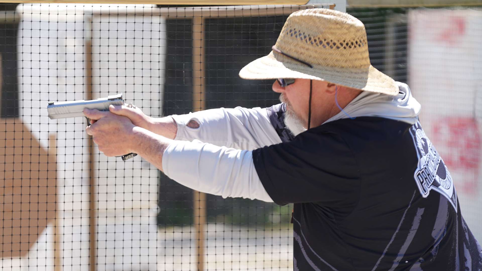 USPSA competitor wearing a straw sun hat, sunglasses, and a black-and-white practical shooting jersey extends a stainless steel competition pistol with both hands