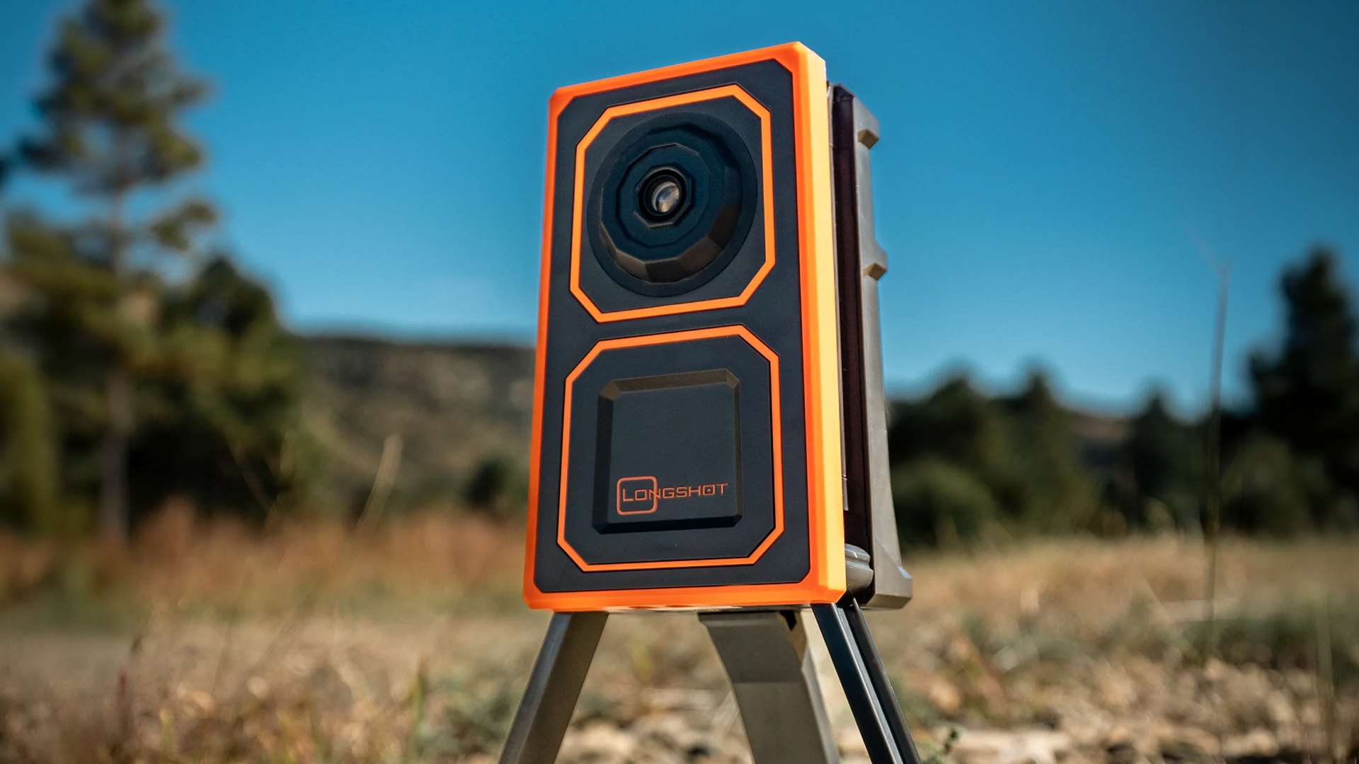The Longshot Ranger target camera, a rectangular black-and-orange unit with a lens up top and the Longshot logo below, mounted on a low tripod in a grassy field with pine trees and hills in the background
