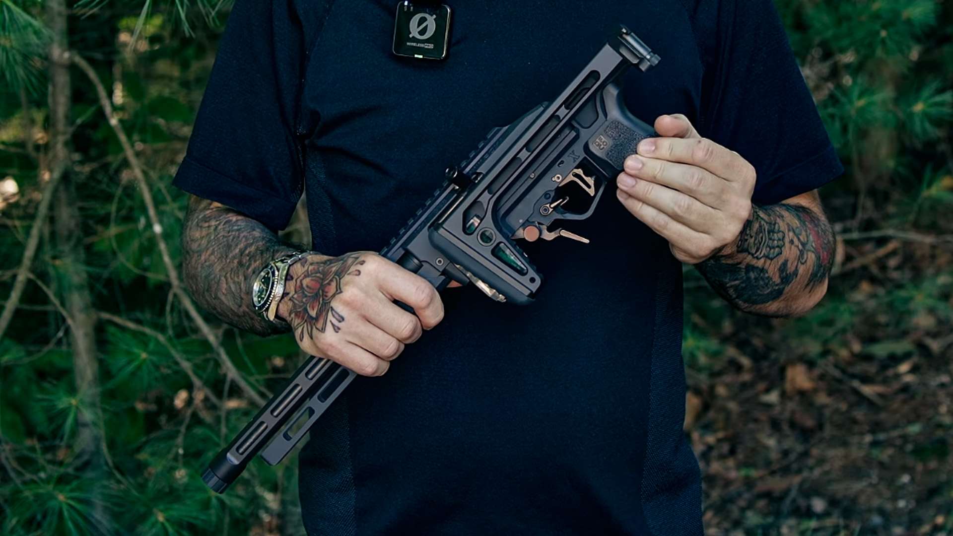 A man holds a Tandemkross ADX SBR with its stock folded alongside the receiver, showing the short M-LOK barrel, textured grip and bronze-colored trigger and controls against a backdrop of pine trees